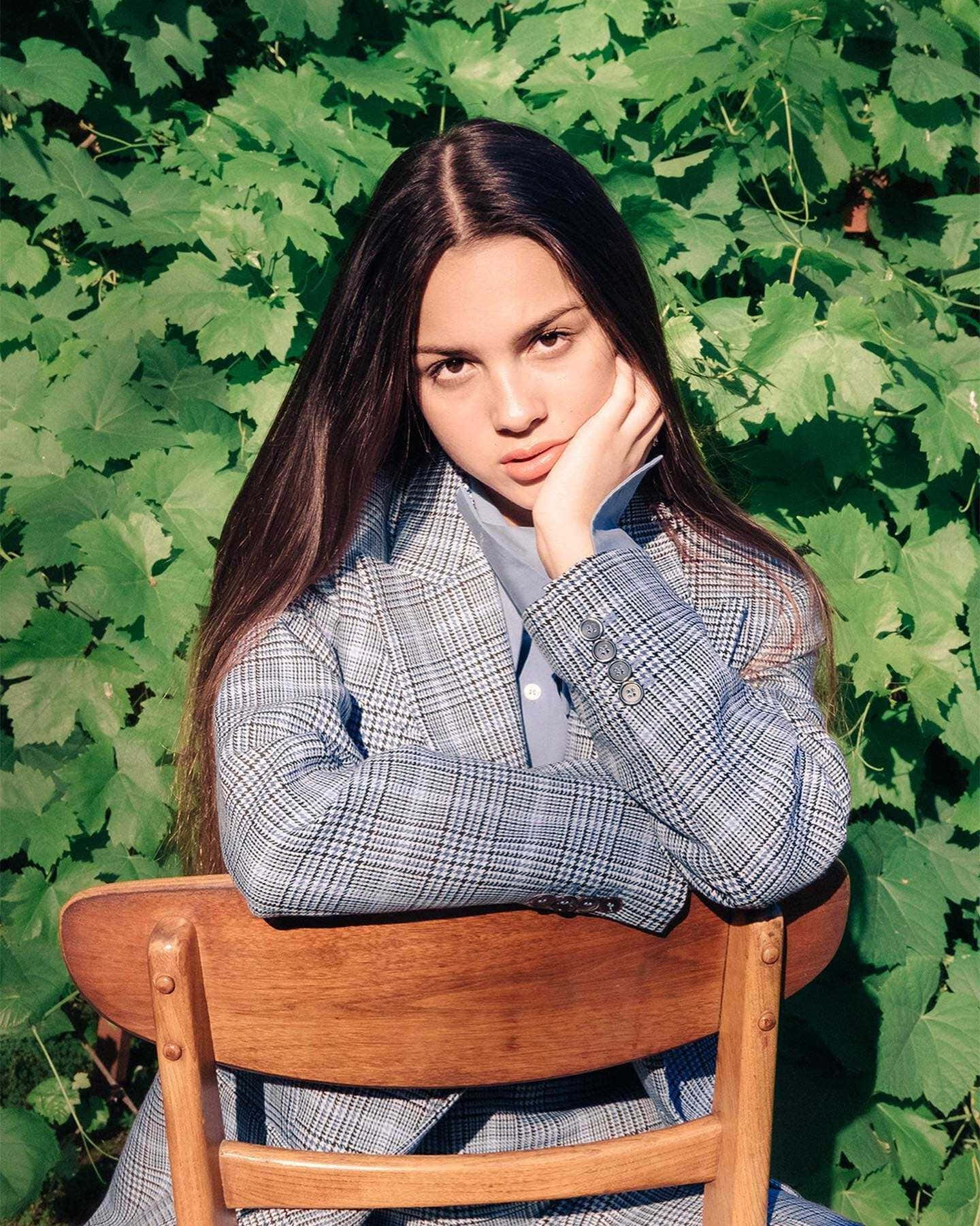 Olivia Rodrigo 桌布 壁紙 — 一位長髮深色嘅年輕女性坐喺木椅上，背景係茂密嘅綠色葉牆，直視鏡頭，表情若有所思。