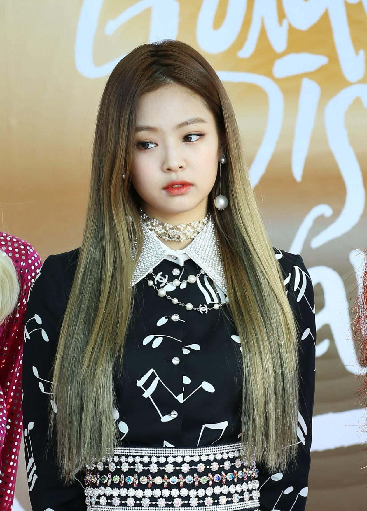 Jennie Kim 桌布 壁紙 — 一位長髮漸層染色的年輕女性，身穿黑色服裝，飾有白色音符和閃亮配飾，背景為金色，帶有白色字樣。