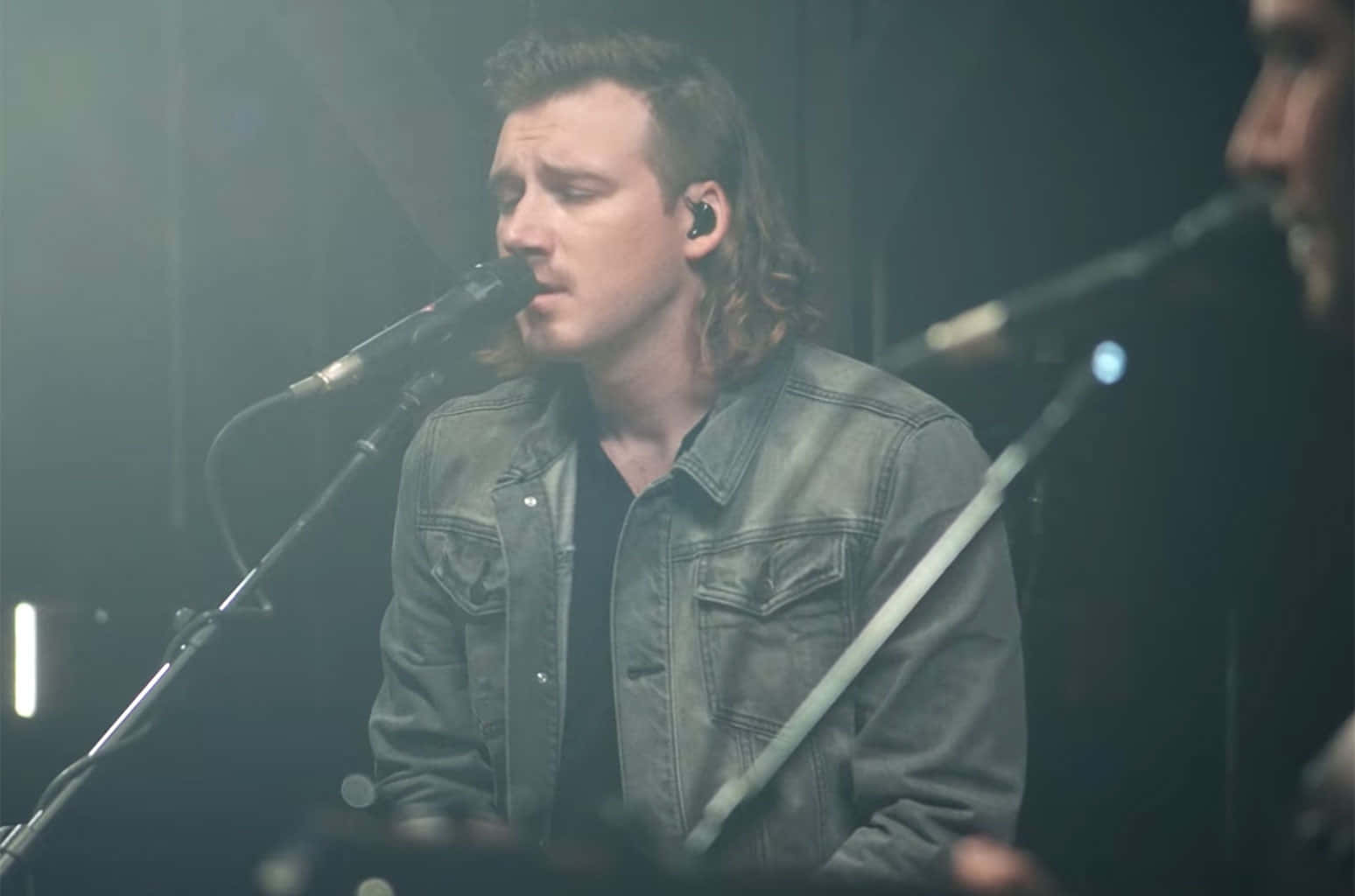 Morgan Wallen 桌布 壁紙 — 一位長髮男歌手在昏暗的舞台燈光下，對著麥克風激情演唱。