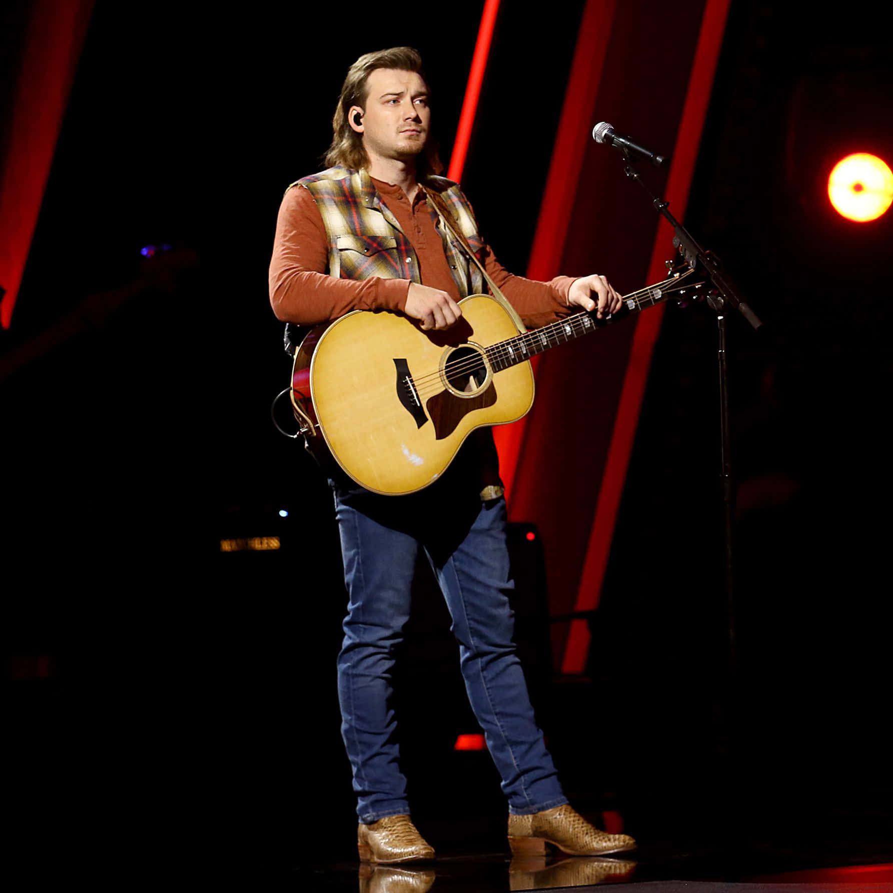 Morgan Wallen 桌布 壁紙 — 一位長髮男音樂家在昏暗的舞台上，在紅色聚光燈下彈奏木結他。