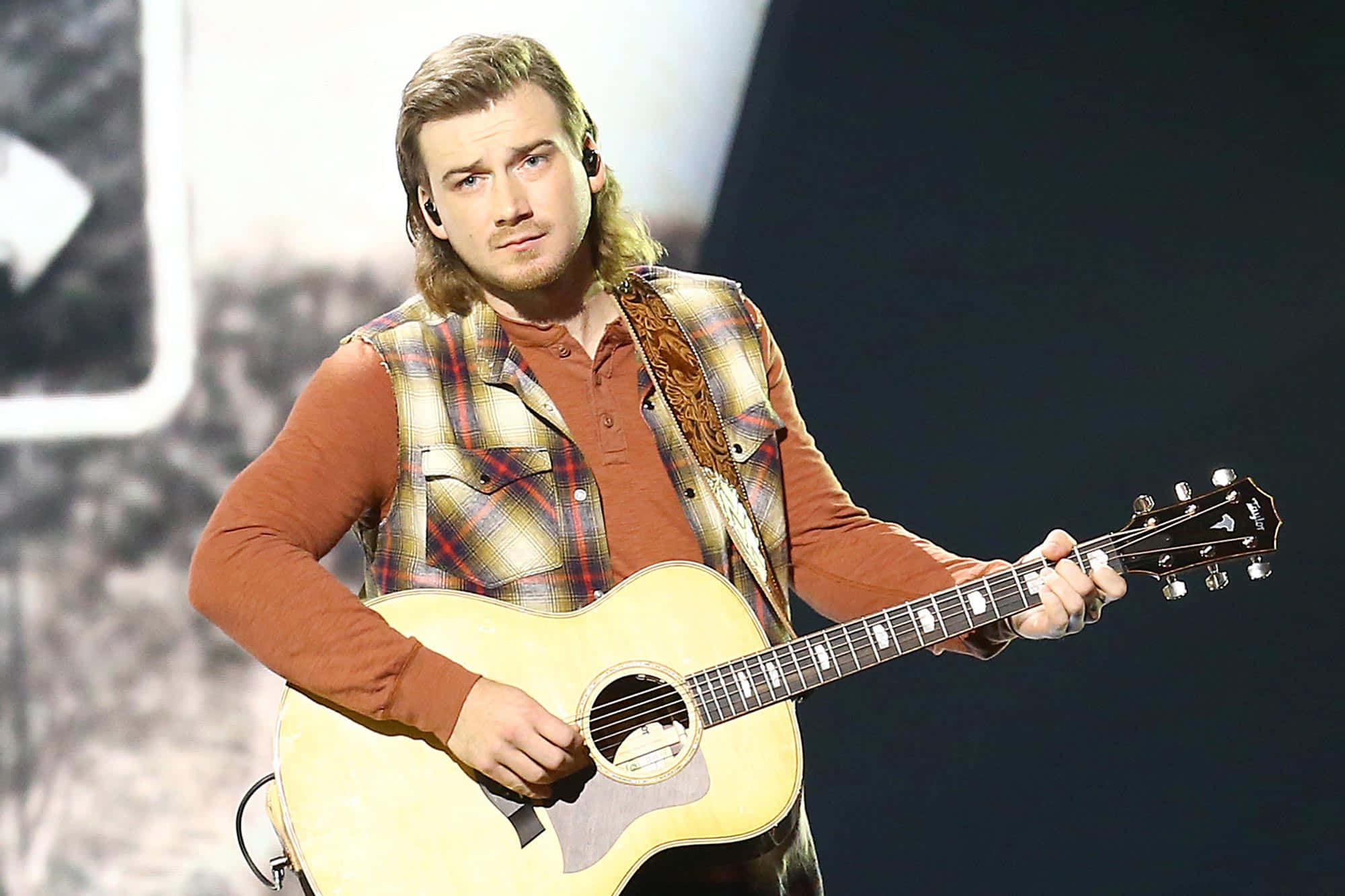 Morgan Wallen 桌布 壁紙 — 一位長髮男音樂家在燈光昏暗的舞台上彈奏原聲結他，身穿銹色襯衫外搭格仔背心。
