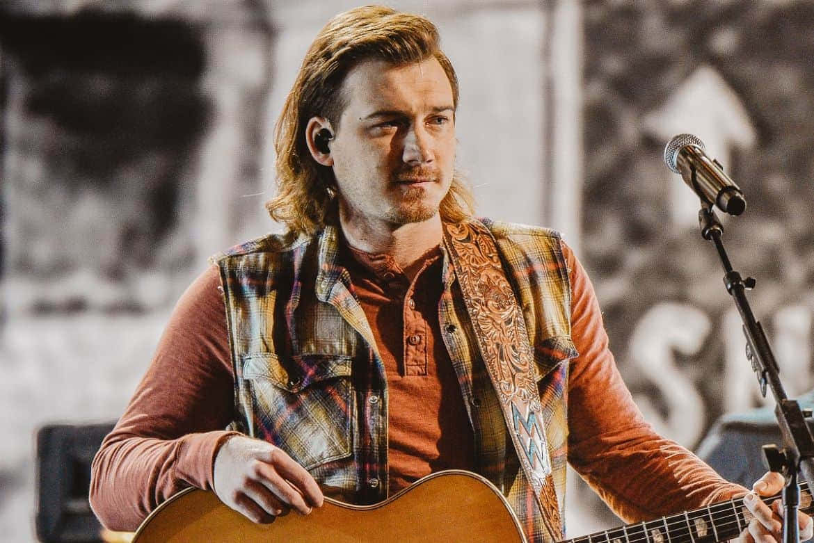 Morgan Wallen 桌布 壁紙 — 一位長髮音樂家在溫暖的舞台燈光下彈奏木結他。