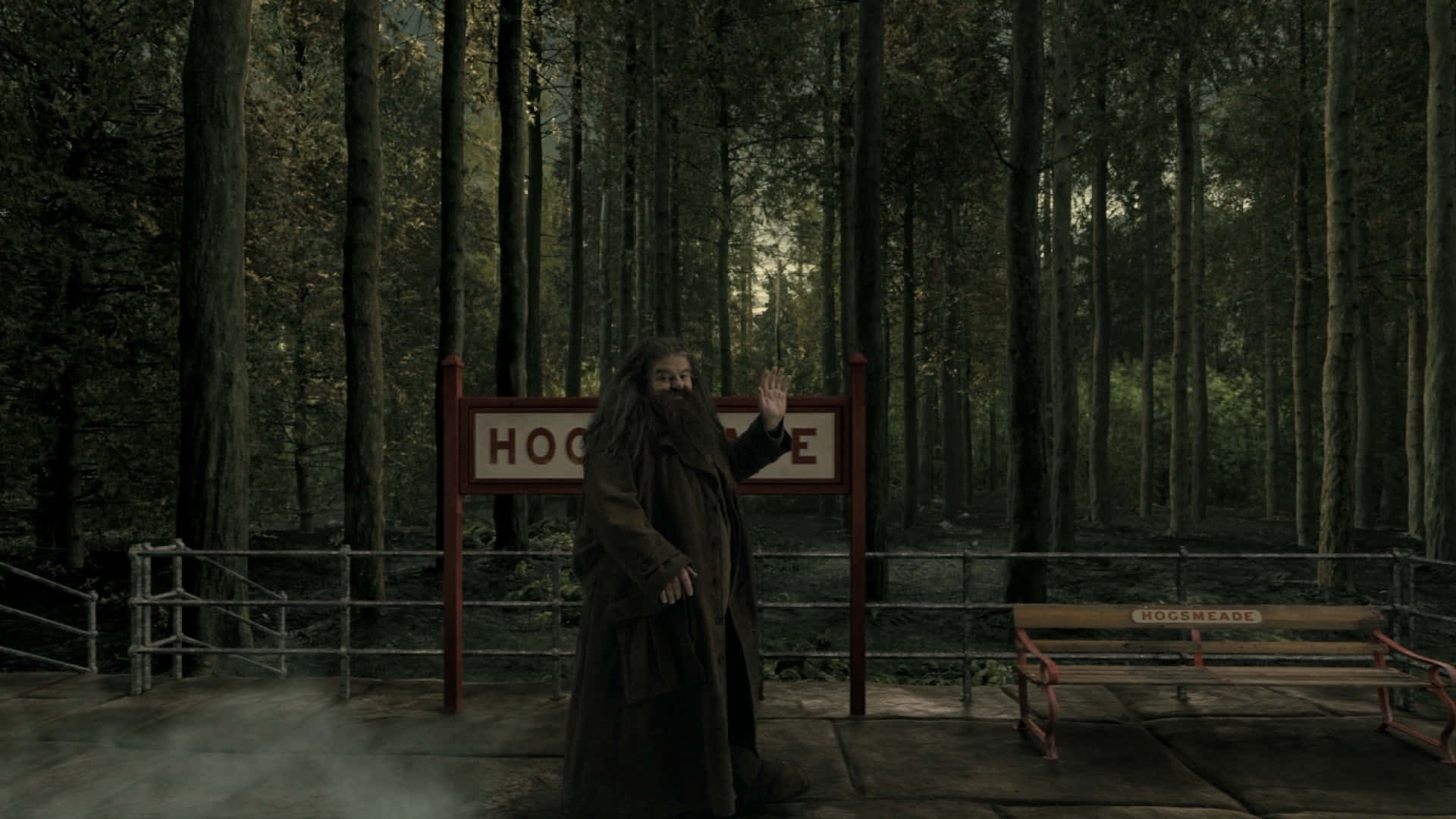 Hogsmeade 桌布 壁紙 — 一位長鬍鬚的巫師在霧氣瀰漫的霍格莫德車站向鏡頭揮手，周圍是高聳的暗色樹木。
