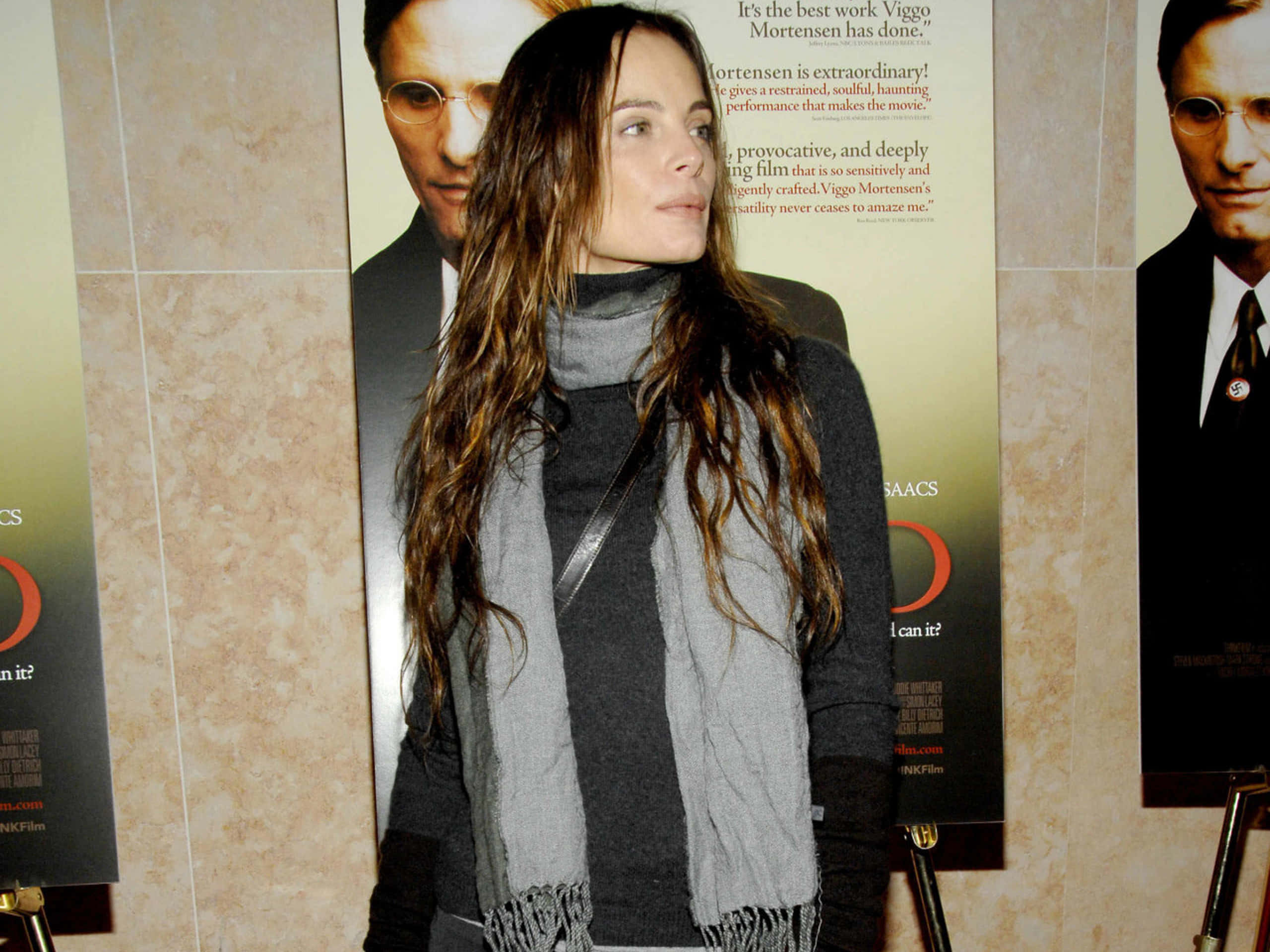 Gabrielle Anwar 桌布 壁紙 — 一位長黑髮、戴灰色圍巾的女性站在鋪瓷磚的走廊裡，旁邊是電影海報。