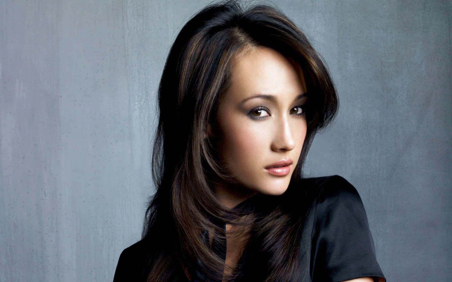 maggie q 桌布 壁紙 — 一位長黑髮、穿黑色上衣的女性，在紋理灰色背景前溫柔凝視。