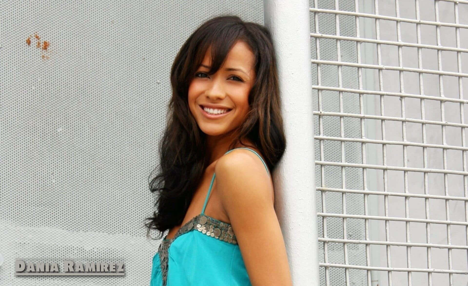 Dania Ramirez 桌布 壁紙 — 一位長黑髮微笑的女子靠在白色格柵圍欄旁的紋理灰牆上。