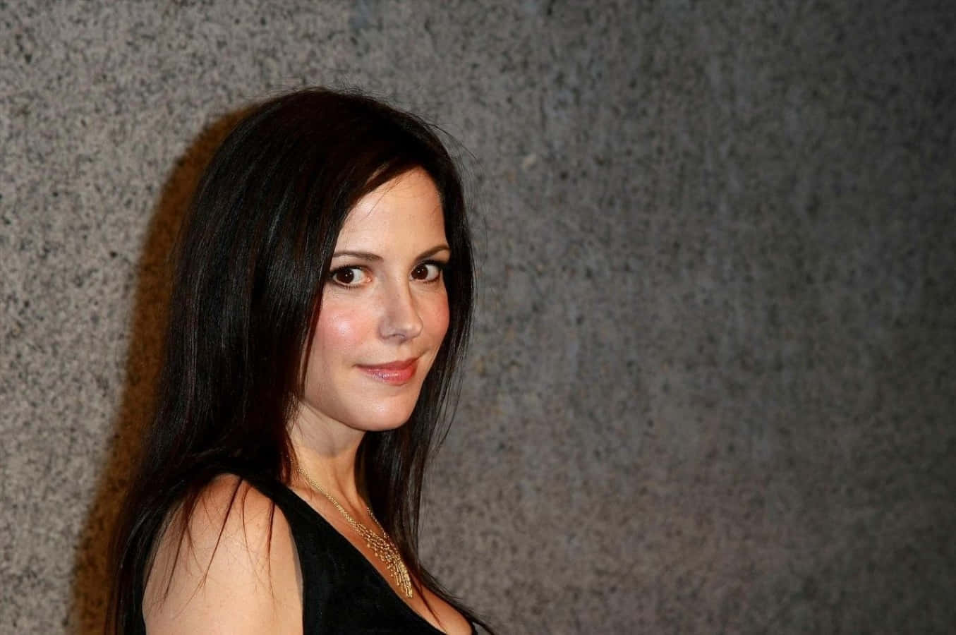 mary louise parker 桌布 壁紙 — 一位長黑髮的女性，在紋理灰色牆壁前，穿著黑色上衣和金色項鍊，溫柔地微笑。