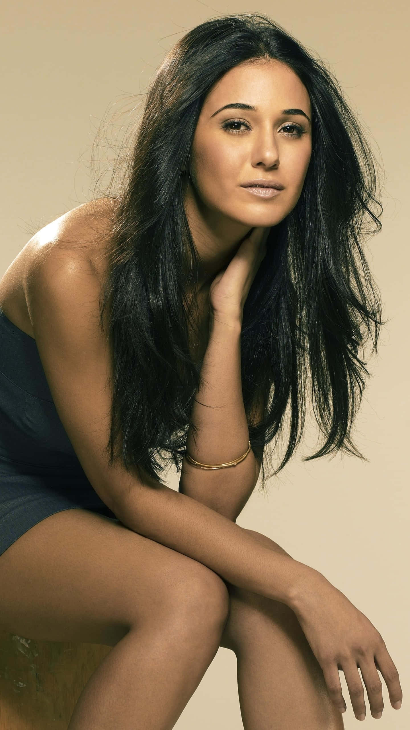 Emmanuelle Chriqui 桌布 壁紙 — 一位長黑髮的女性優雅地坐在攝影棚中，手放在脖子上，背景是柔和的米色。