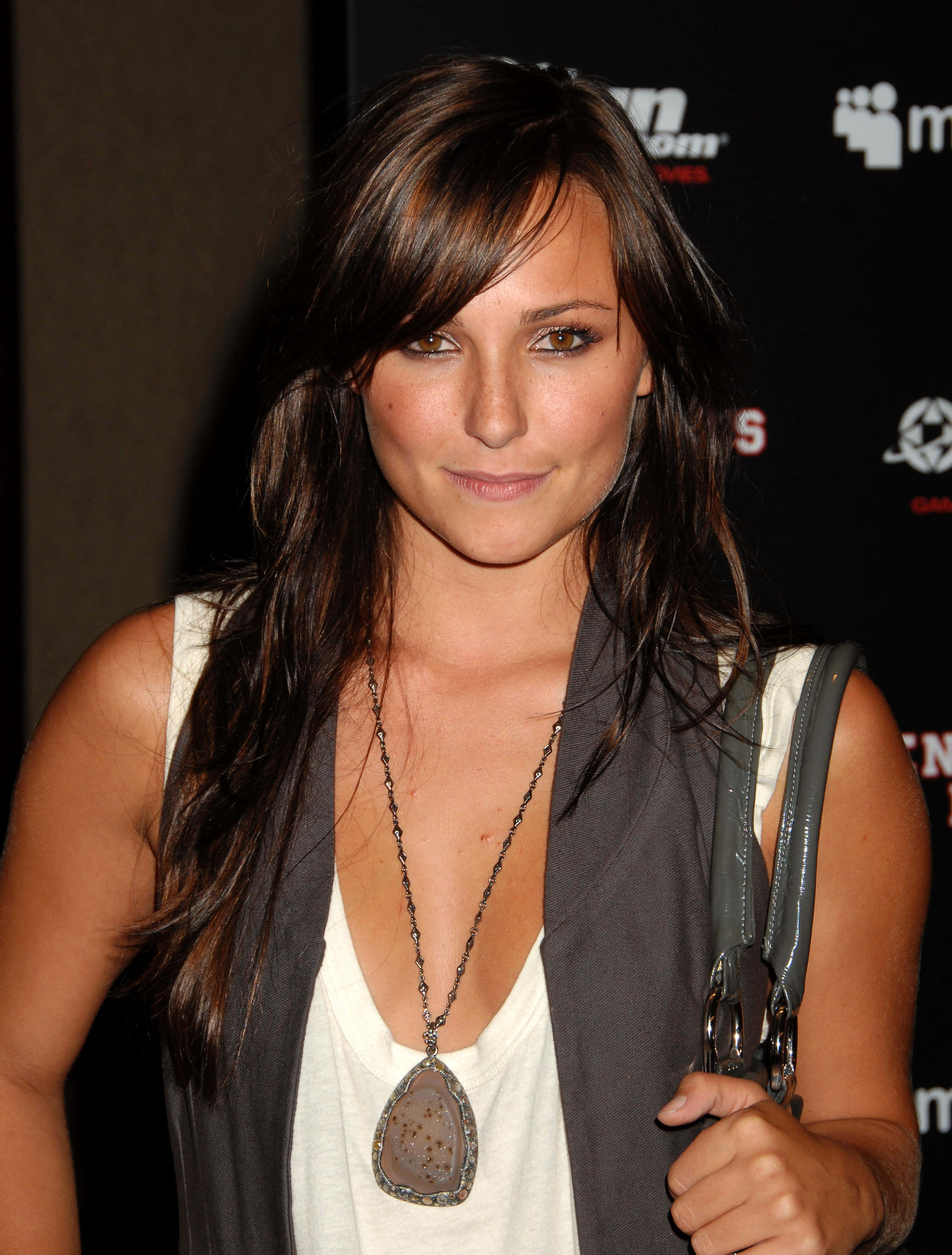 Briana Evigan 桌布 壁紙 — 一位長黑髮的女性在品牌活動中穿著白色上衣和灰色背心，溫柔地微笑。
