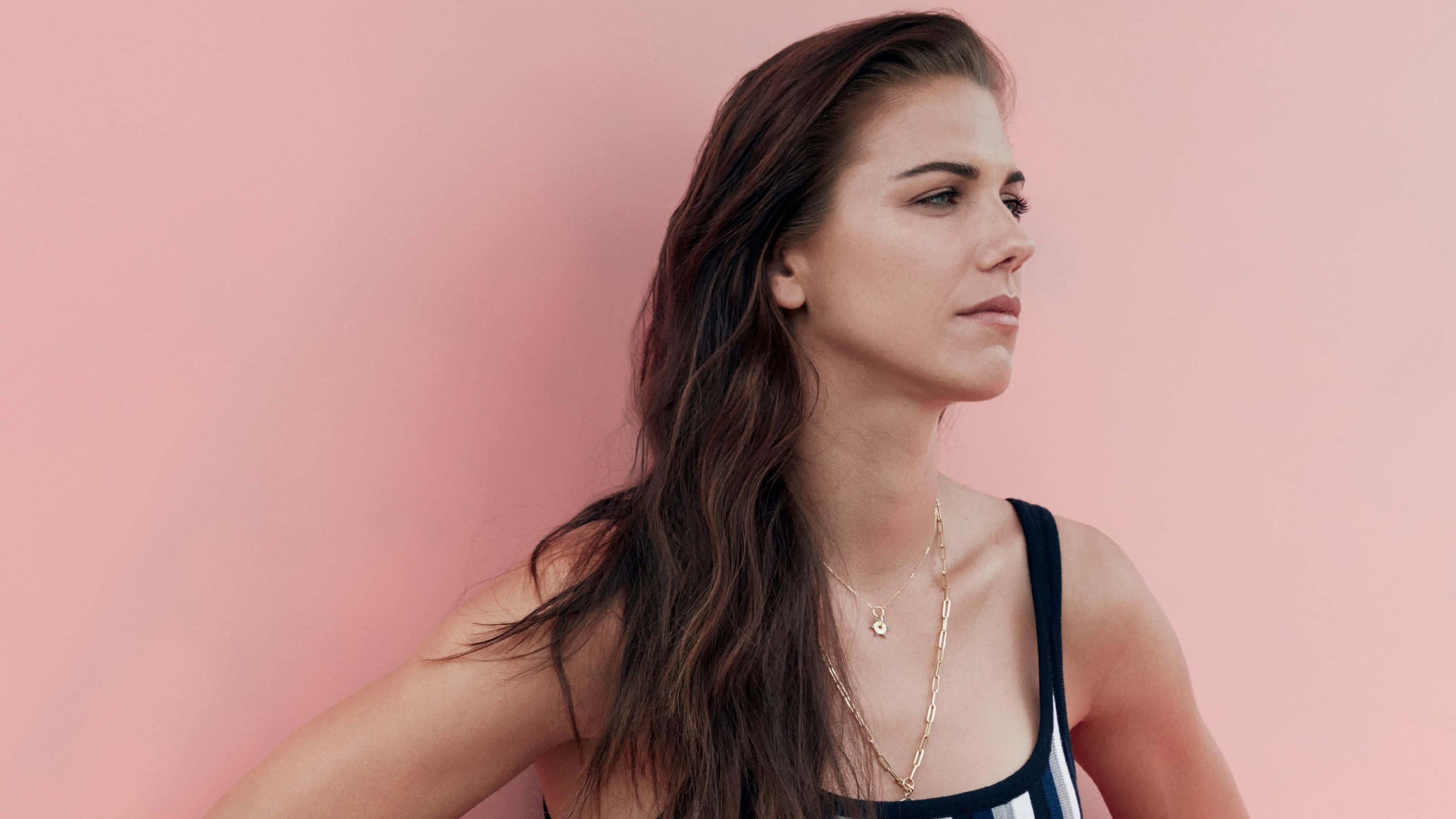 Alex Morgan 桌布 壁紙 — 一位長黑髮的女性在柔和的粉紅色工作室背景前，穿著黑白上衣，若有所思地凝視著。