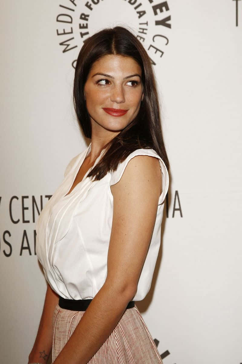 Genevieve Padalecki 桌布 壁紙 — 一位長黑髮的女性在活動中微笑，身穿白色無袖上衣和條紋裙。