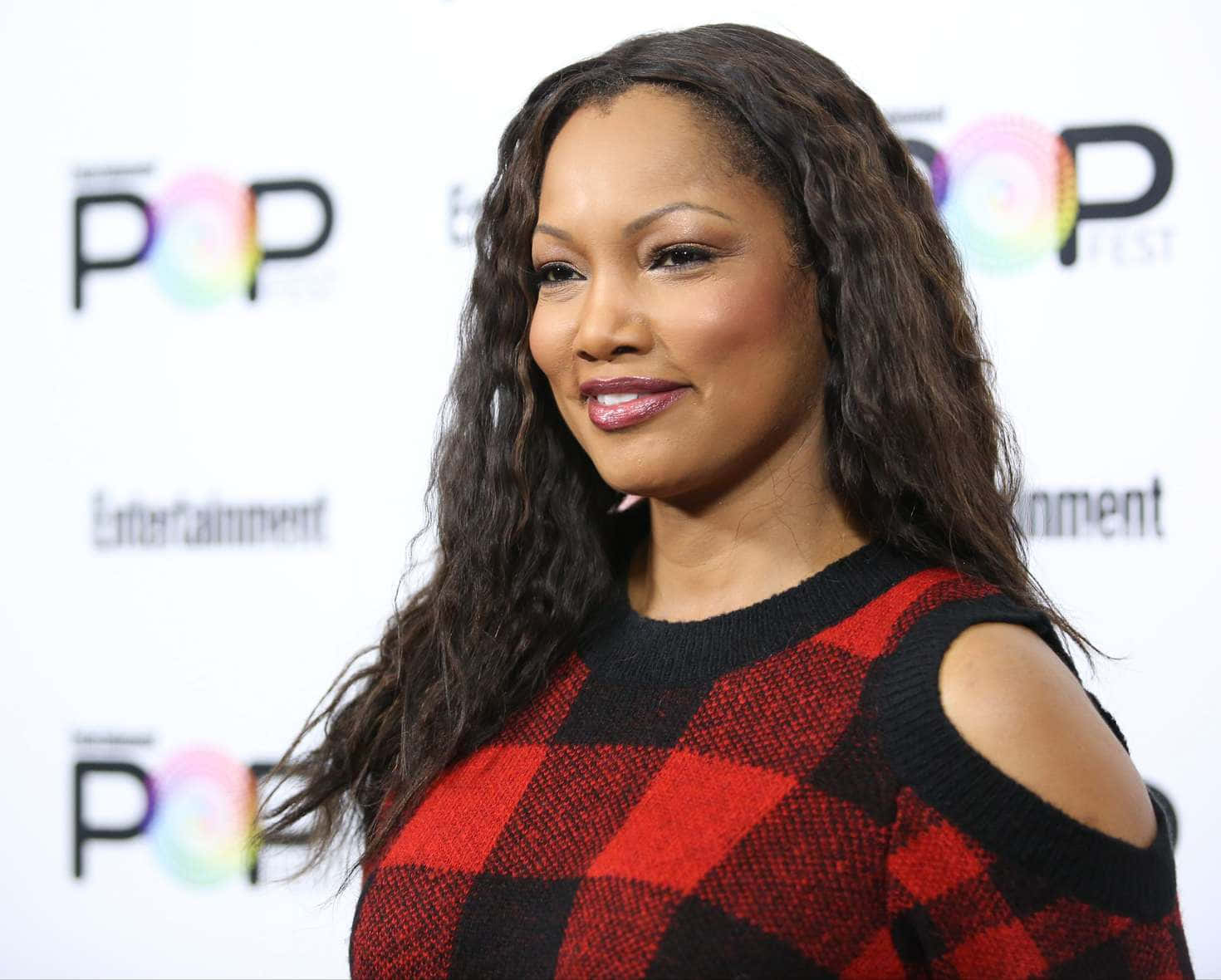 Garcelle Beauvais 桌布 壁紙 — 一位長黑髮的女性在流行音樂節活動中穿著紅黑格紋毛衣，溫暖地微笑。