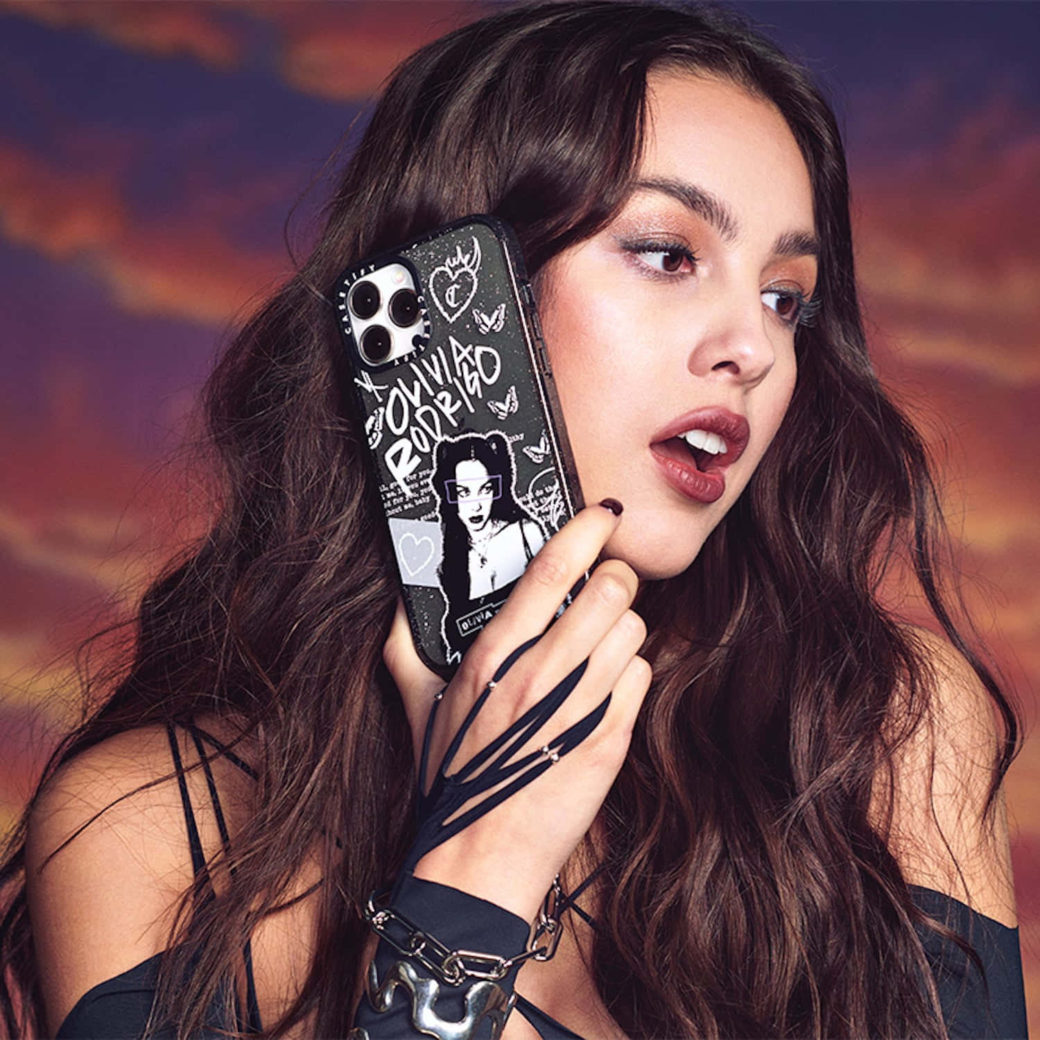 Olivia Rodrigo 桌布 壁紙 — 一位長黑髮的女性手持帶有圖案保護殼的手機，背景是火紅的日落天空。
