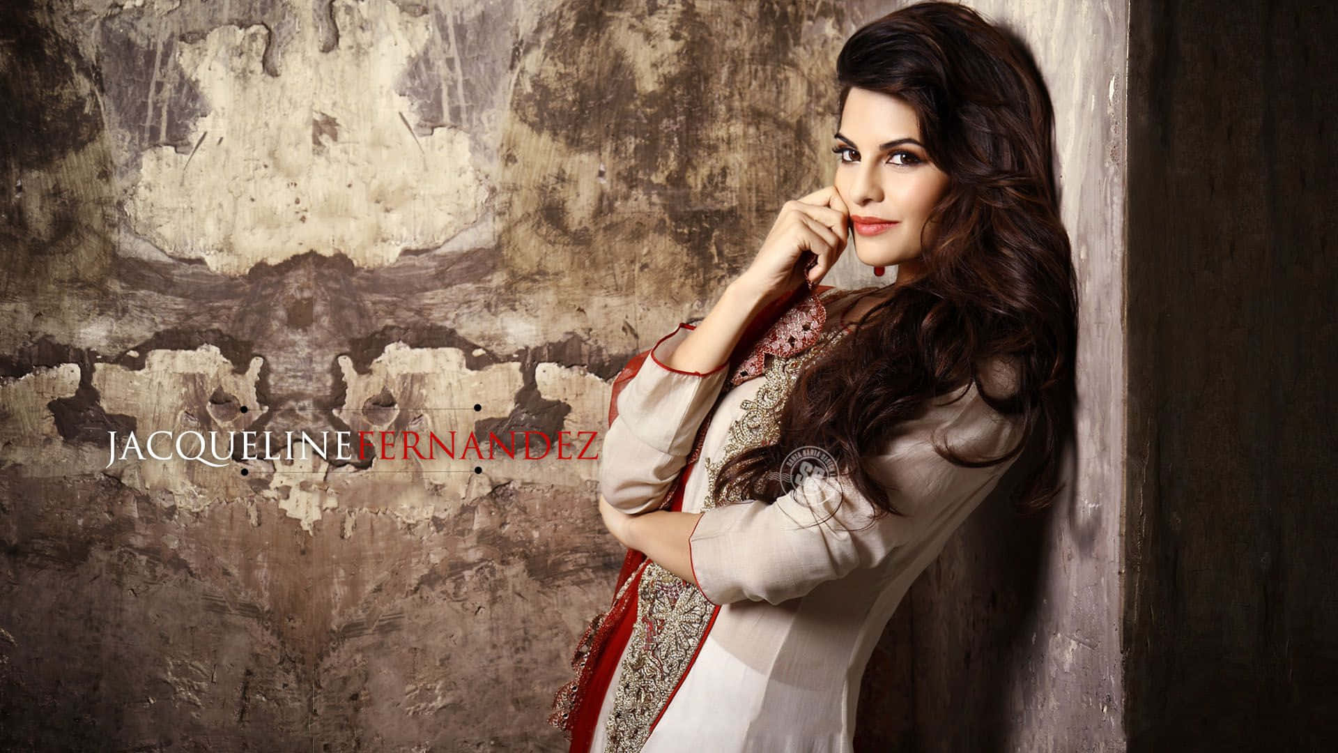 Jacqueline Fernandez 桌布 壁紙 — 一位長黑髮的女性靠著風化的牆壁，身穿米色和紅色刺繡服裝，自信地凝視鏡頭。