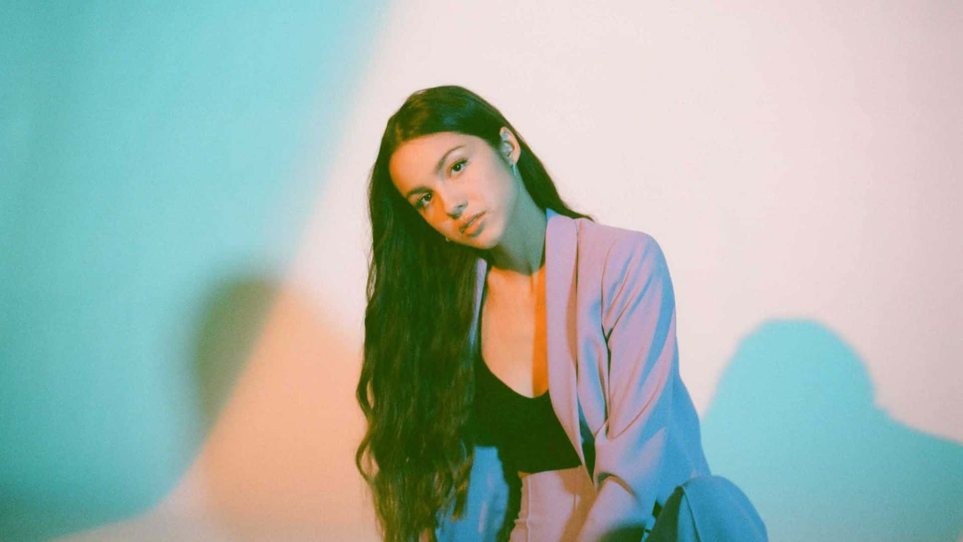 Olivia Rodrigo 桌布 壁紙 — 一位長黑髮的年輕女性在柔和燈光的牆前擺姿勢，牆壁沐浴在冷藍色和暖粉色的光中。