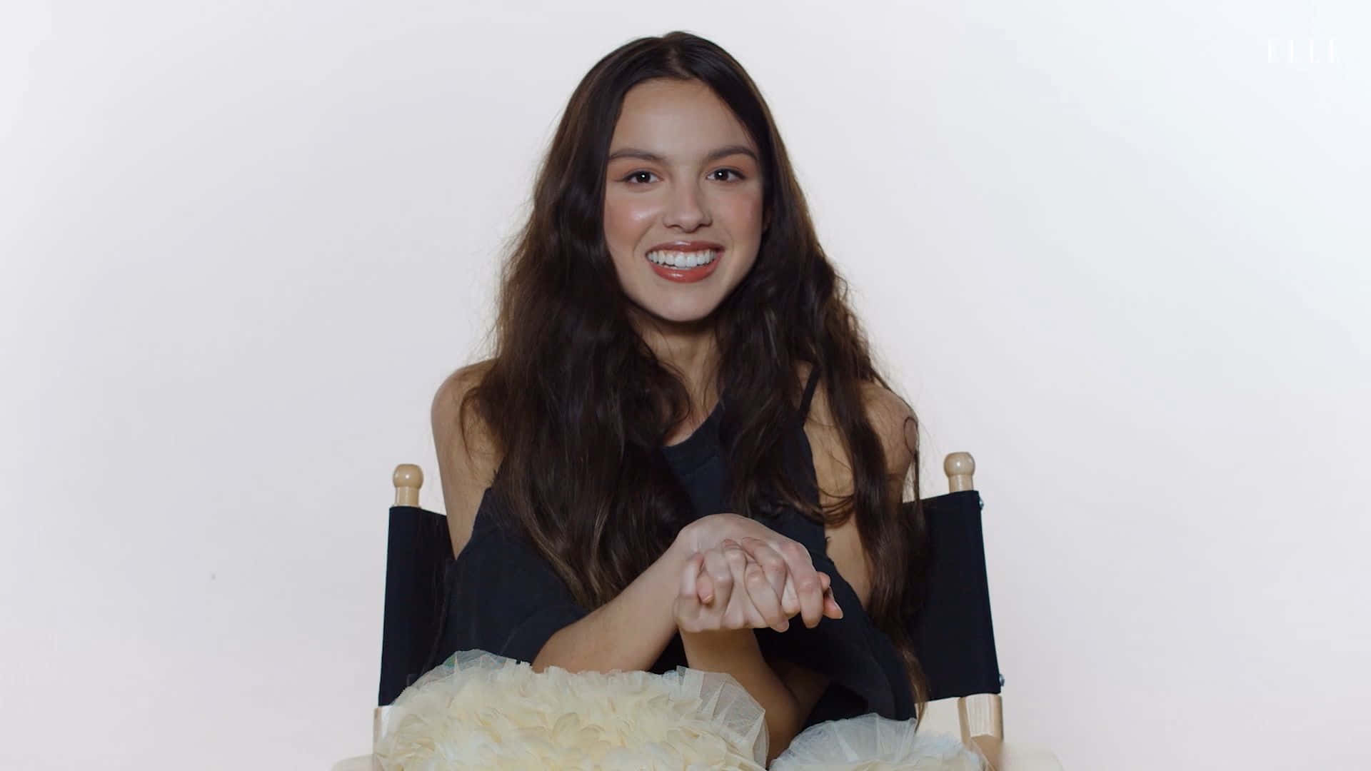 Olivia Rodrigo 桌布 壁紙 — 一位長黑髮的年輕女性坐在導演椅上，對著純白背景燦爛微笑。