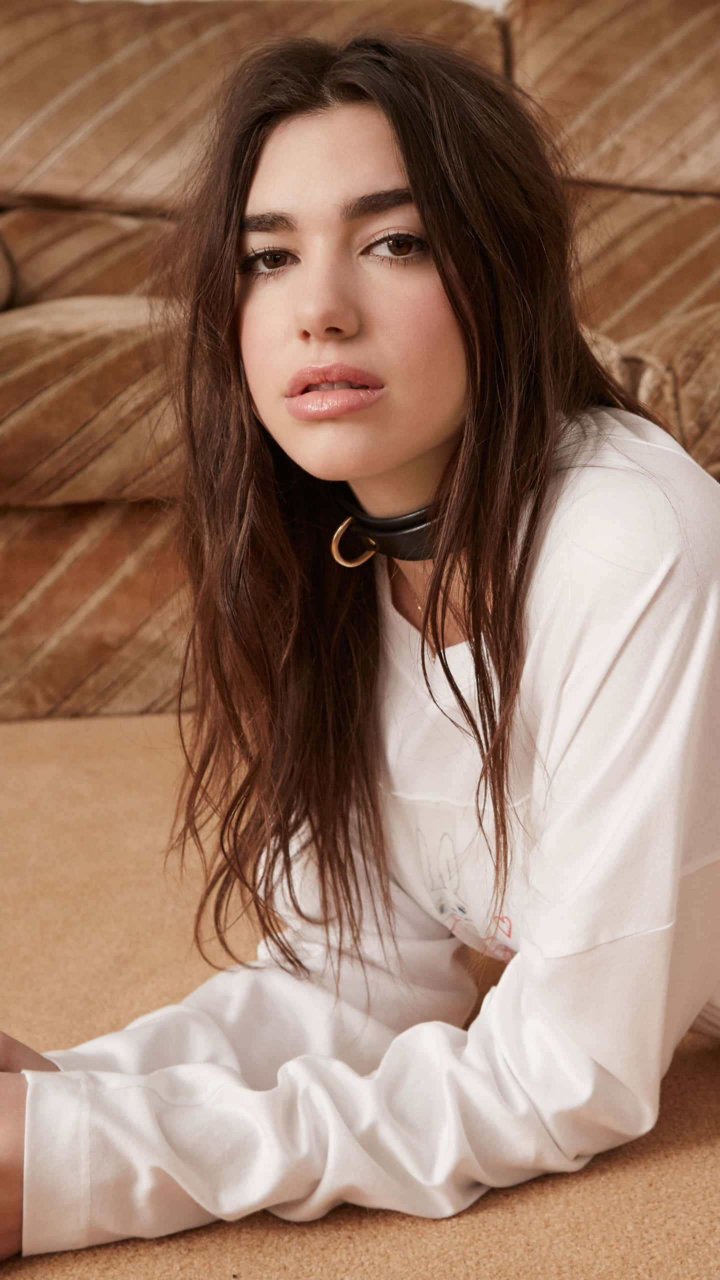 Dua Lipa 桌布 壁紙 — 一位長黑髮的年輕女性躺在有紋理的棕色沙發上，穿著白色襯衫和黑色頸鏈。