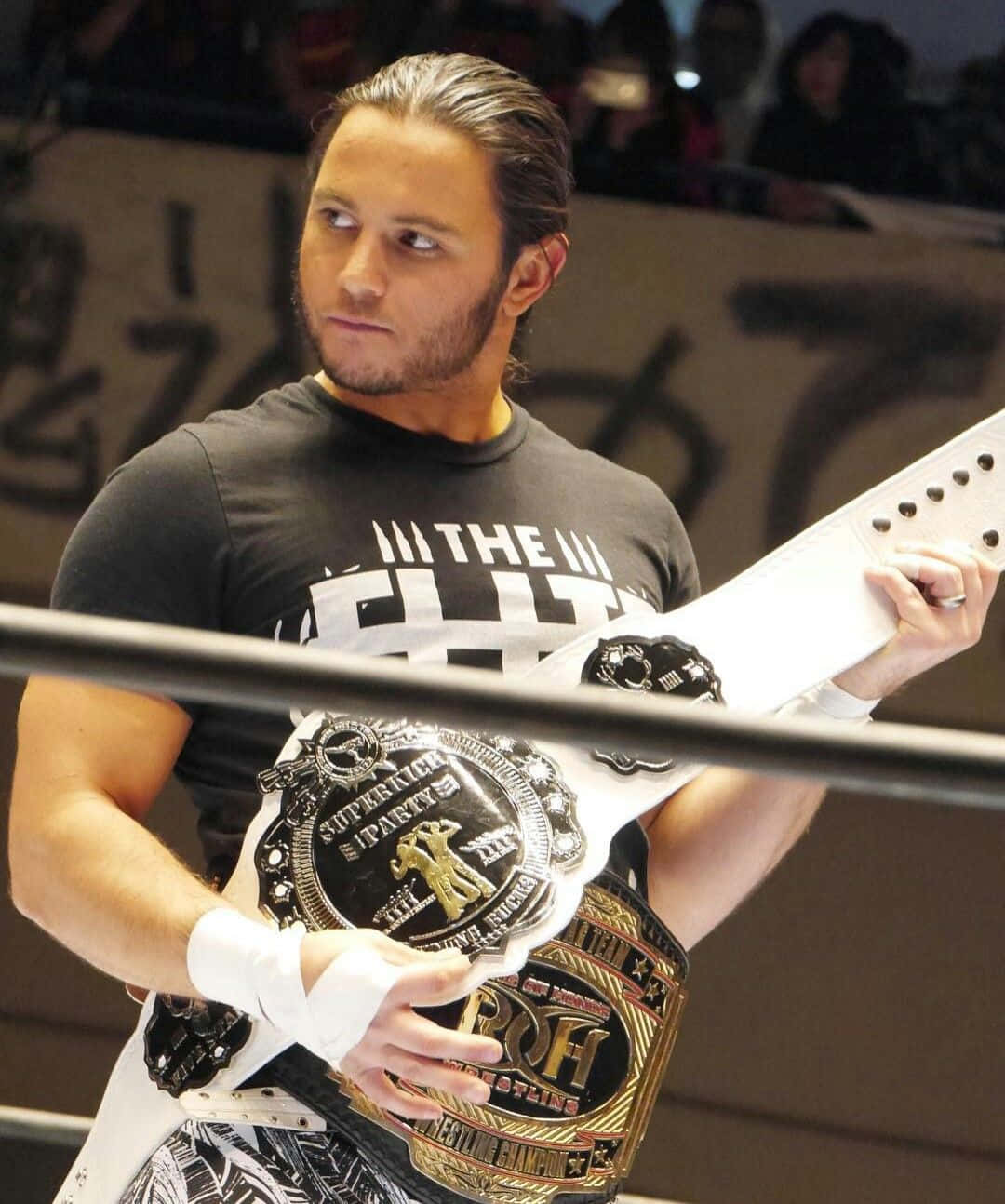 American Professional Wrestler Matt Jackson Superkick Party Belt 壁紙 — 一位長黑髮的摔角手在明亮的場館燈光下於擂台上高舉冠軍腰帶。