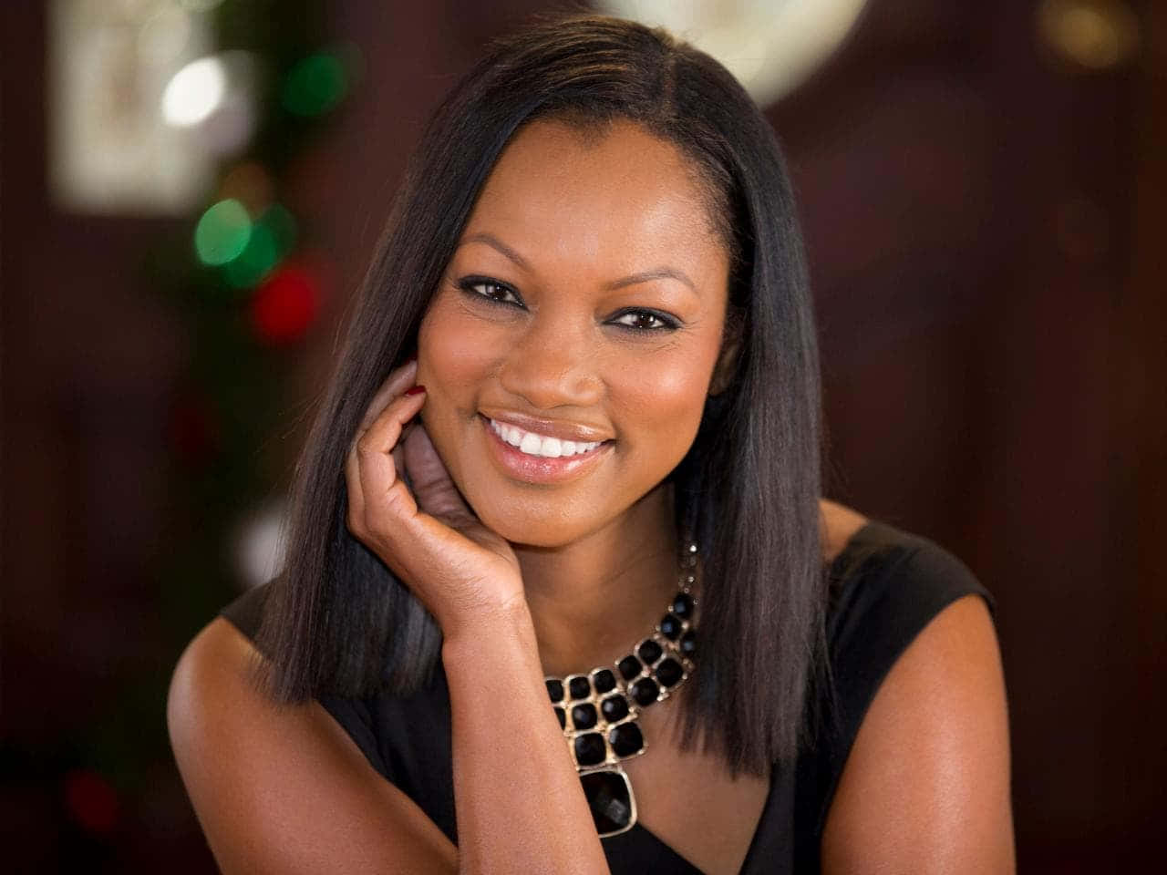 Garcelle Beauvais 桌布 壁紙 — 一位長黑髮的燦爛女性，在柔和模糊的節日背景前，將下巴擱在手上溫暖地微笑。