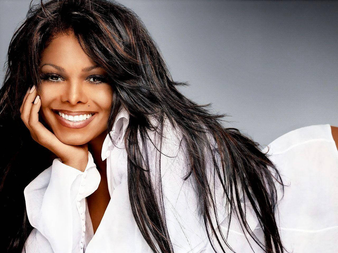 R&B Junkie Janet Jackson 壁紙 — 一位長黑髮的燦爛女性，在柔和的灰色背景下，將下巴擱在手上，溫暖地微笑。