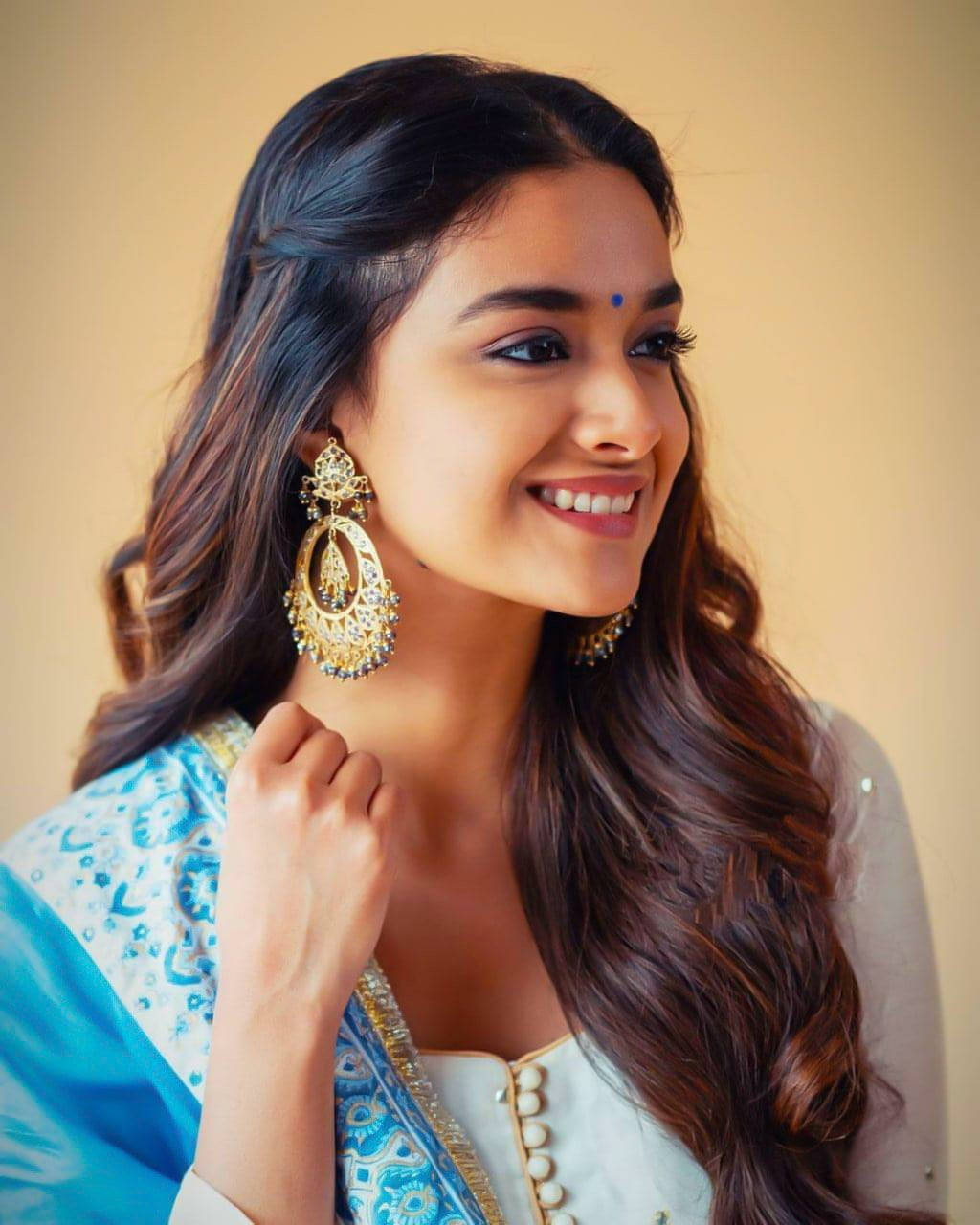 Tamil Actors HD 桌布 壁紙 — 一位長黑髮的燦爛女性，在柔和的米色背景前，戴著華麗的金耳環和藍色圖案服裝，溫暖地微笑。