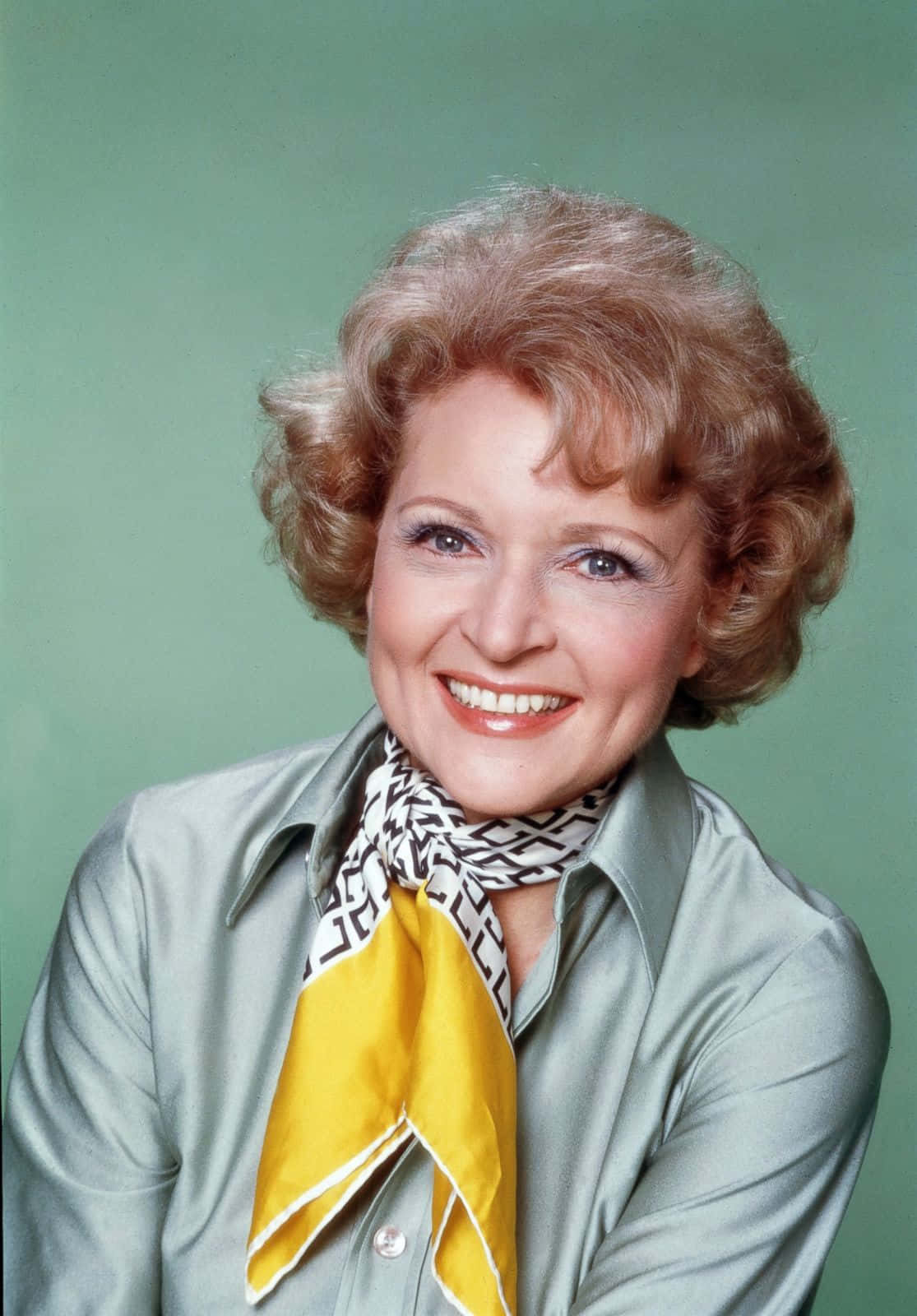 Betty White 桌布 壁紙 — 一位開心的女性，擁有捲曲金髮，穿著銀色襯衫和黃色圖案圍巾，在綠色背景前溫暖地微笑。