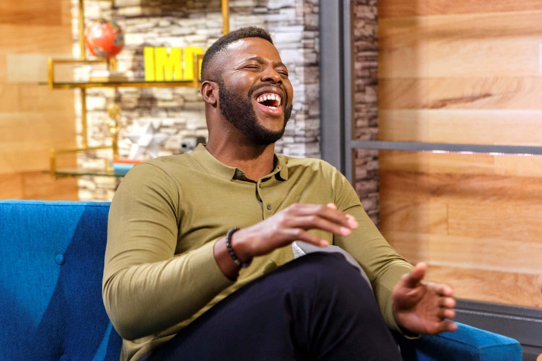 Winston Duke 桌布 壁紙 — 一位開心的黑人男子坐在現代工作室的藍色沙發上開懷大笑。