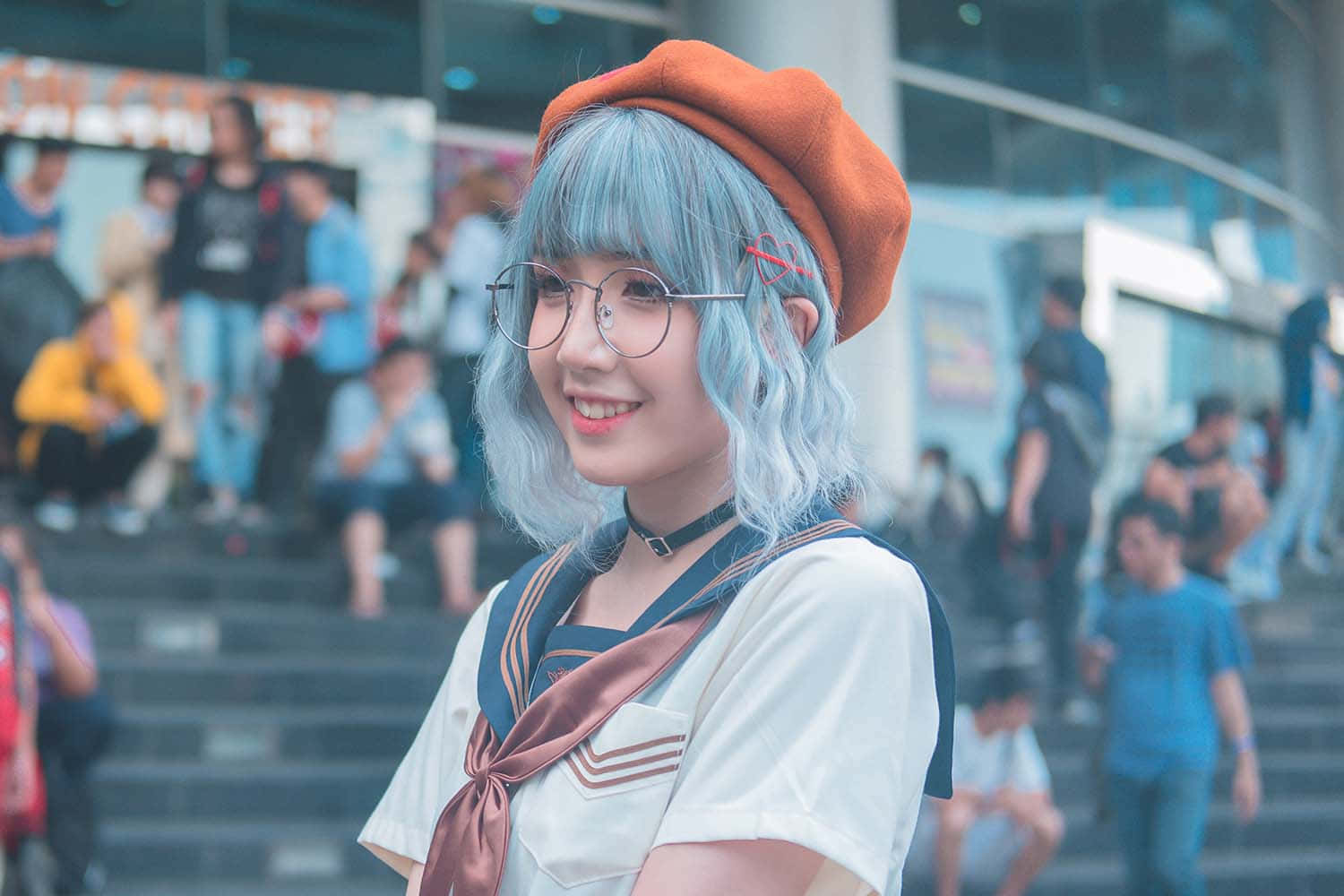 Otaku 桌布 壁紙 — 一位開朗的動漫風格女孩，藍色頭髮戴著橙色貝雷帽，在繁華的城市廣場對著鏡頭微笑。