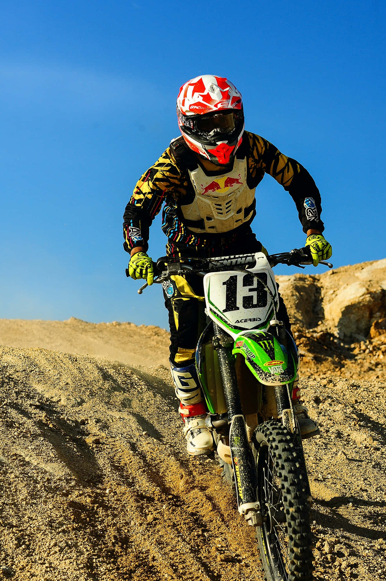 Motocross 桌布 壁紙 — 一位電單車越野賽車手戴著紅白頭盔和黃色手套，在晴朗的藍天下傾身進入一個塵土飛揚的泥地跳躍。