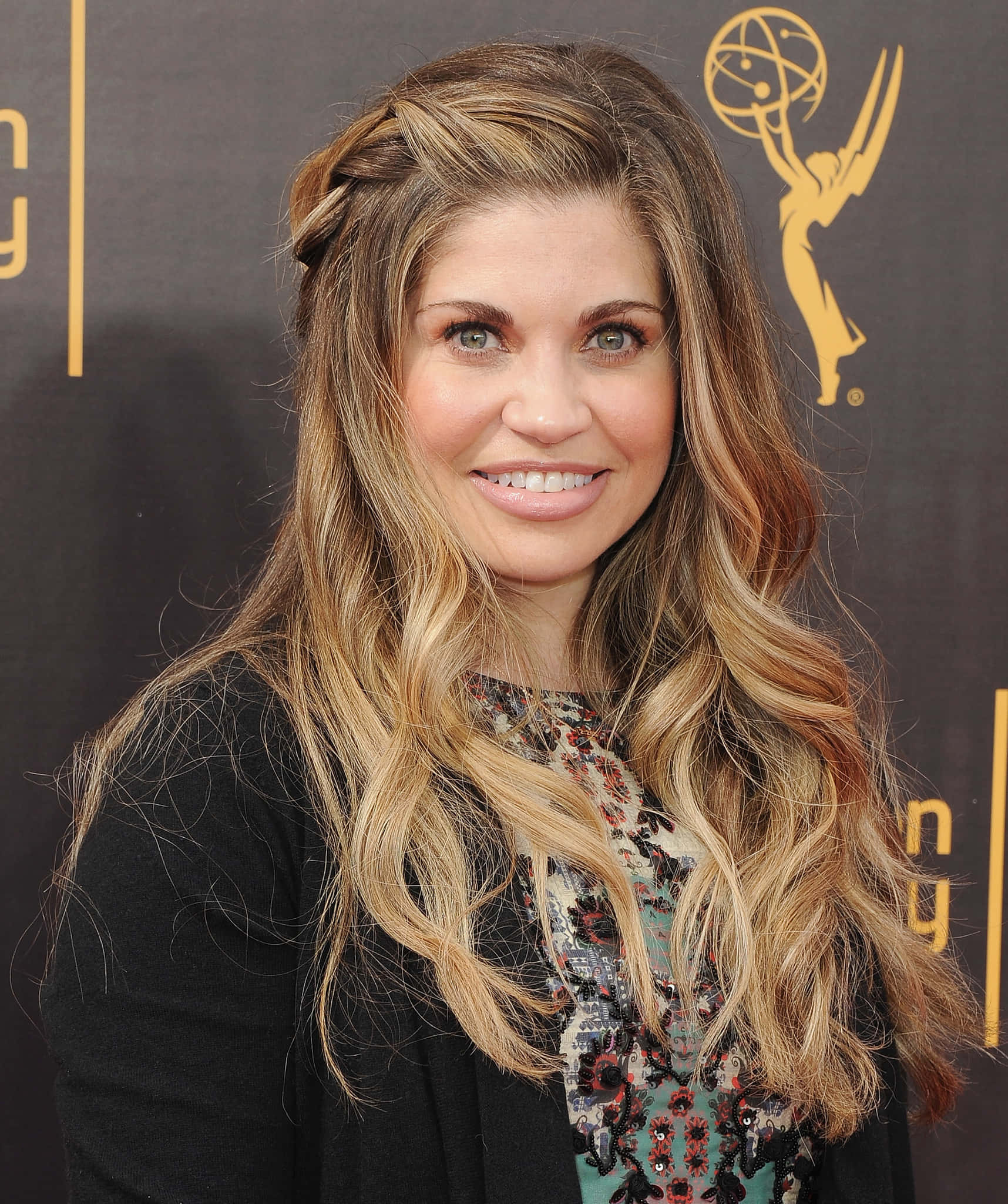 Danielle Fishel 桌布 壁紙 — 一位面帶微笑、擁有長波浪金髮的女性在艾美獎頒獎典禮上，以帶有金色標誌的深色背景為襯托擺姿勢。