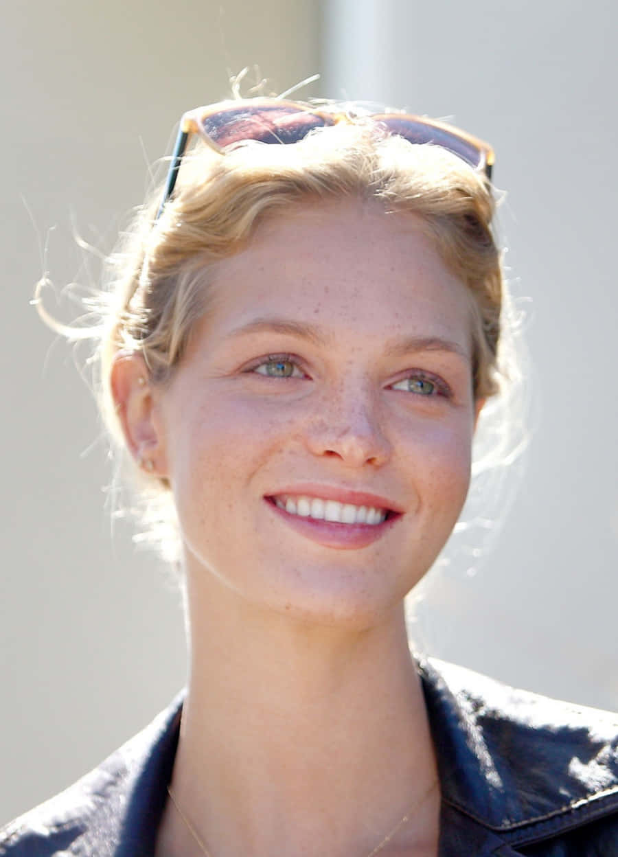 Beautiful Woman 桌布 壁紙 — 一位面帶微笑、有雀斑和金色頭髮的女性，在明亮的背景下，頭上戴著太陽眼鏡，身穿深色外套。