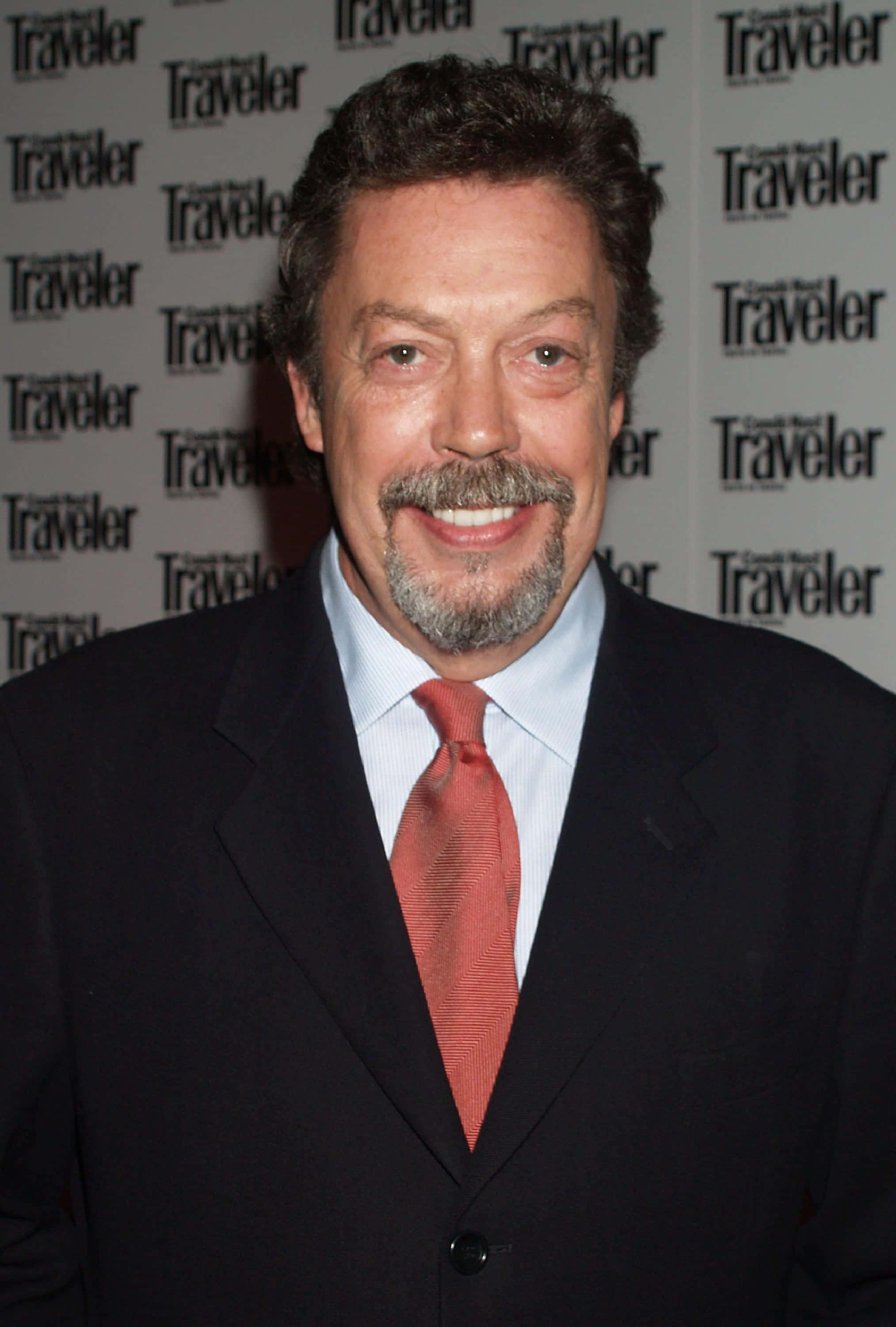 Tim Curry 桌布 壁紙 — 一位面帶微笑、留著灰白鬍鬚和鬍子的男士，穿著深色西裝和紅色領帶，背景是重複的Traveler雜誌圖案。