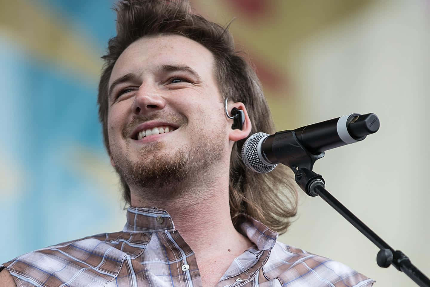 Morgan Wallen 桌布 壁紙 — 一位面帶微笑、留長髮、戴耳機的男音樂家，在明亮的日光下於戶外音樂會對著麥克風唱歌。