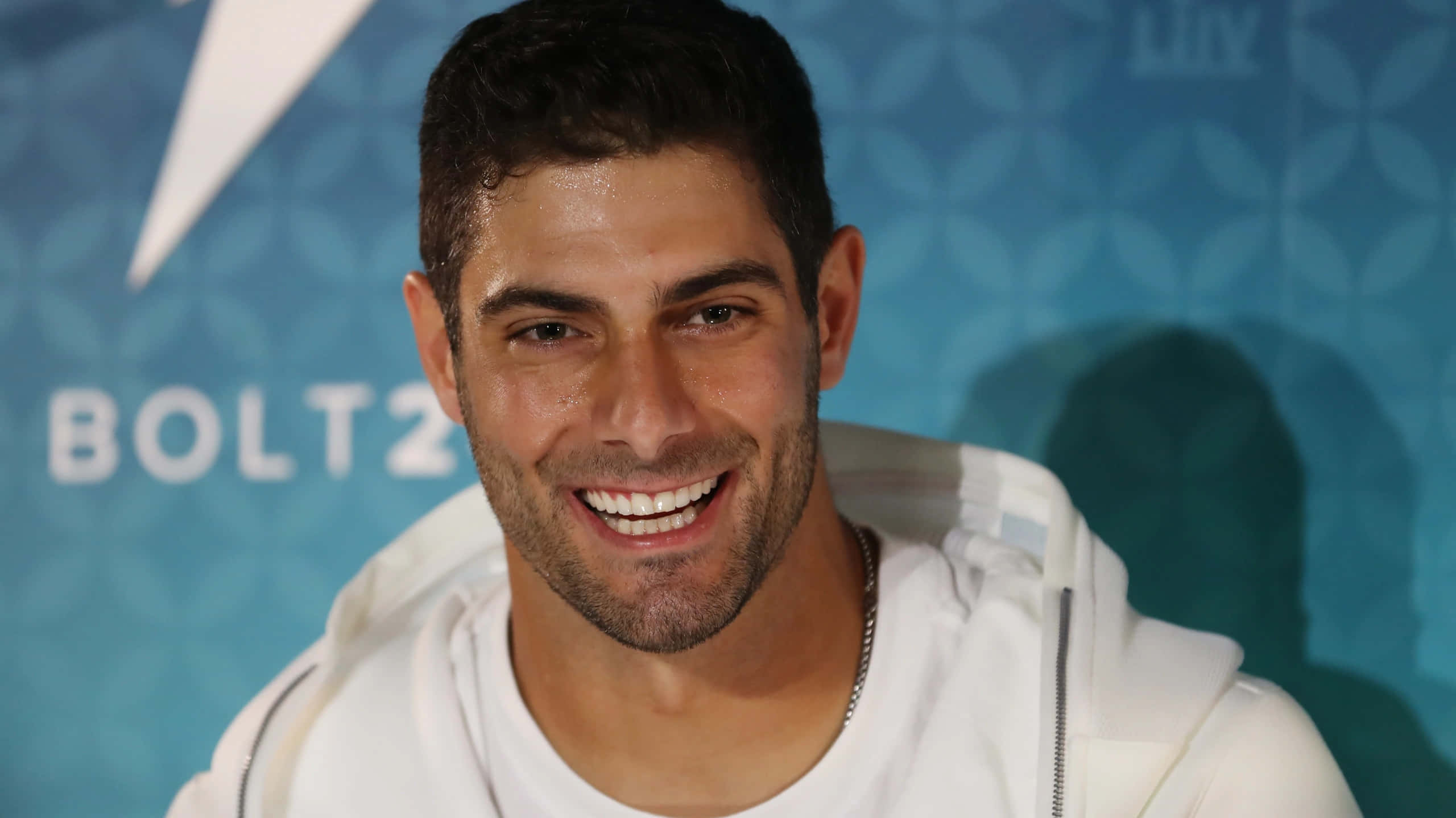 Jimmy Garoppolo 桌布 壁紙 — 一位面帶微笑、短黑髮、鬍渣的男子，身穿白色連帽衫，背景為藍色圖案並帶有 'BOLT2' 文字。