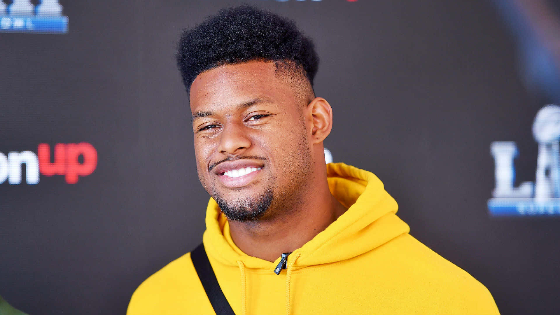 Juju Smith Schuster 桌布 壁紙 — 一位面帶微笑、身穿亮黃色連帽衫的男子在帶有活動標誌的深色背景前擺姿勢。