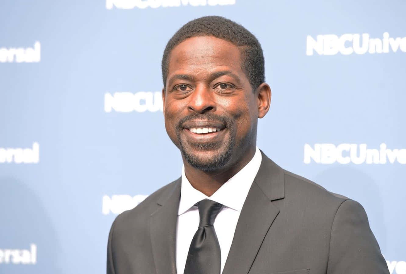 Sterling K Brown 桌布 壁紙 — 一位面帶微笑、身穿深色西裝和領帶的男子，自信地站在藍色NBCUniversal品牌背景前。