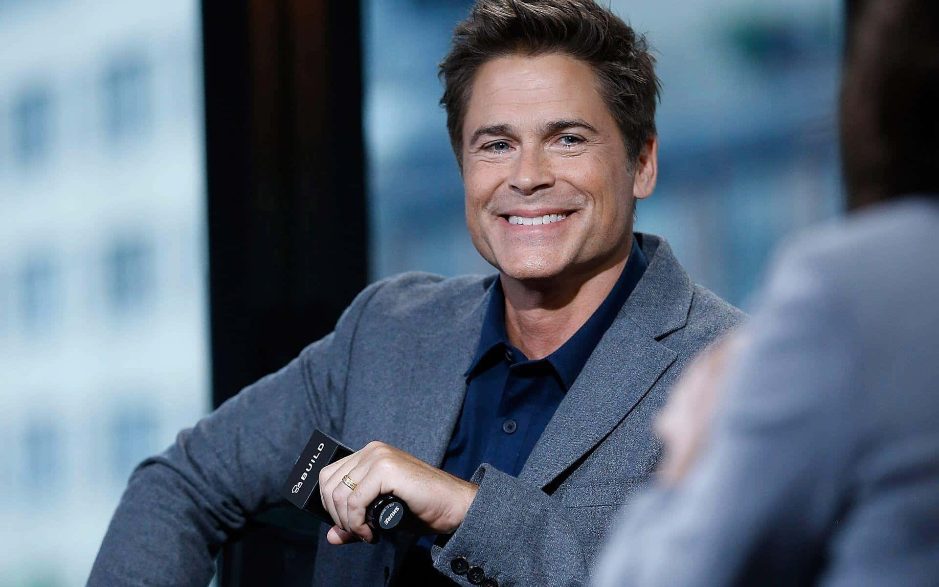 rob lowe 桌布 壁紙 — 一位面帶微笑、身穿灰色西裝外套和深色襯衫的男子，在模糊的城市景觀背景下接受採訪時手持麥克風。