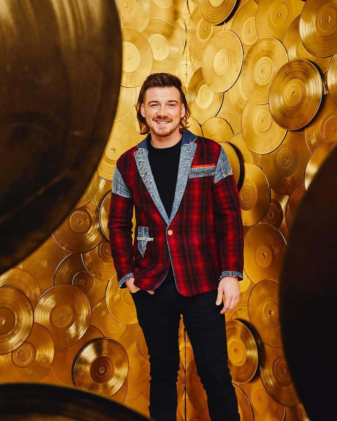 Morgan Wallen 桌布 壁紙 — 一位面帶微笑、身穿紅色格仔外套的男子自信地站在金唱片牆前。
