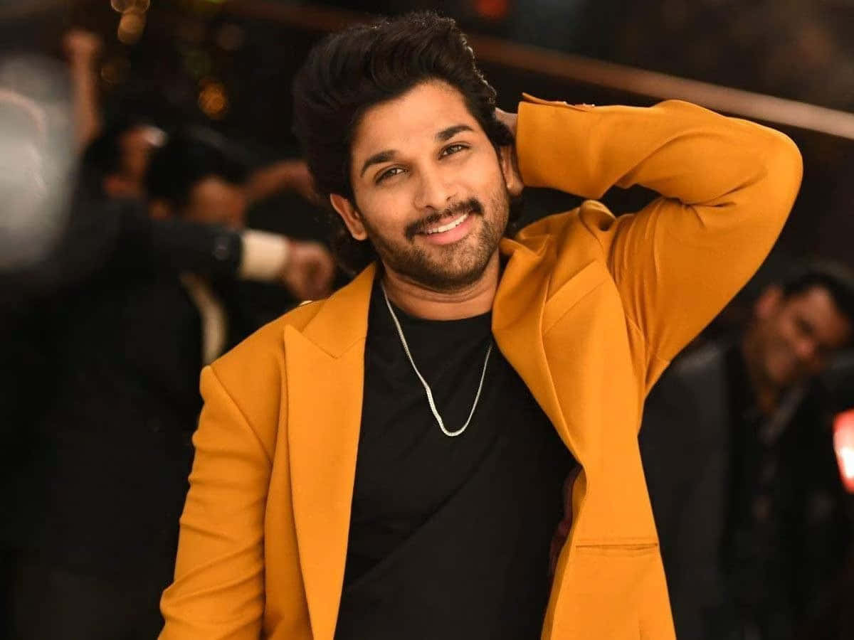 Allu Arjun 桌布 壁紙 — 一位面帶微笑、身穿芥末黃西裝外套的男士，在模糊的派對背景前，自信地將一隻手放在腦後擺姿勢。