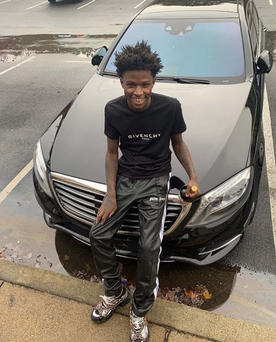 Quando Rondo 桌布 壁紙 — 一位面帶微笑、頭髮豎起的年輕男子靠在一輛停在濕漉漉街道上、滿地落葉的時尚黑色豪華轎車旁。