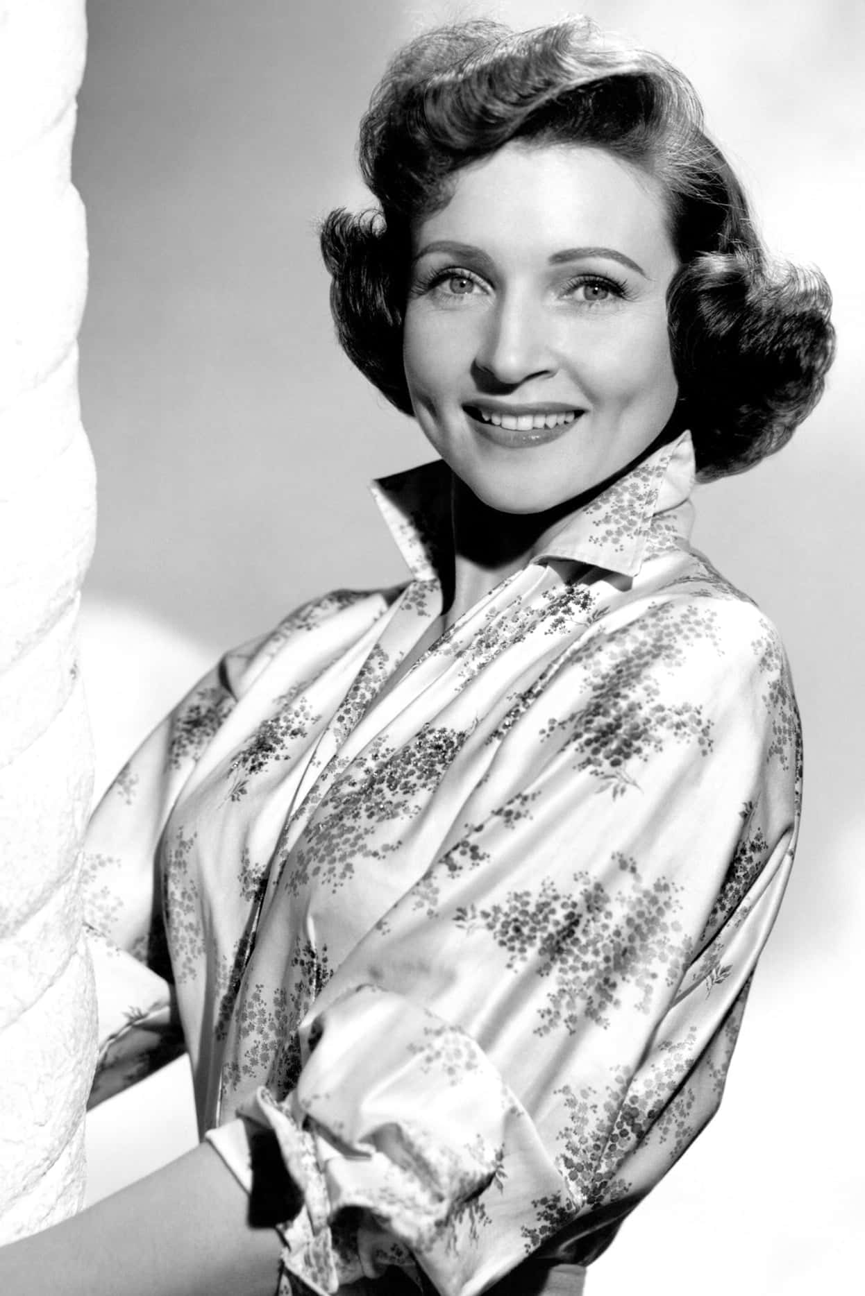Betty White 桌布 壁紙 — 一位面帶微笑、頭髮造型時尚的女性，穿著花卉圖案襯衫，在經典黑白工作室肖像中。