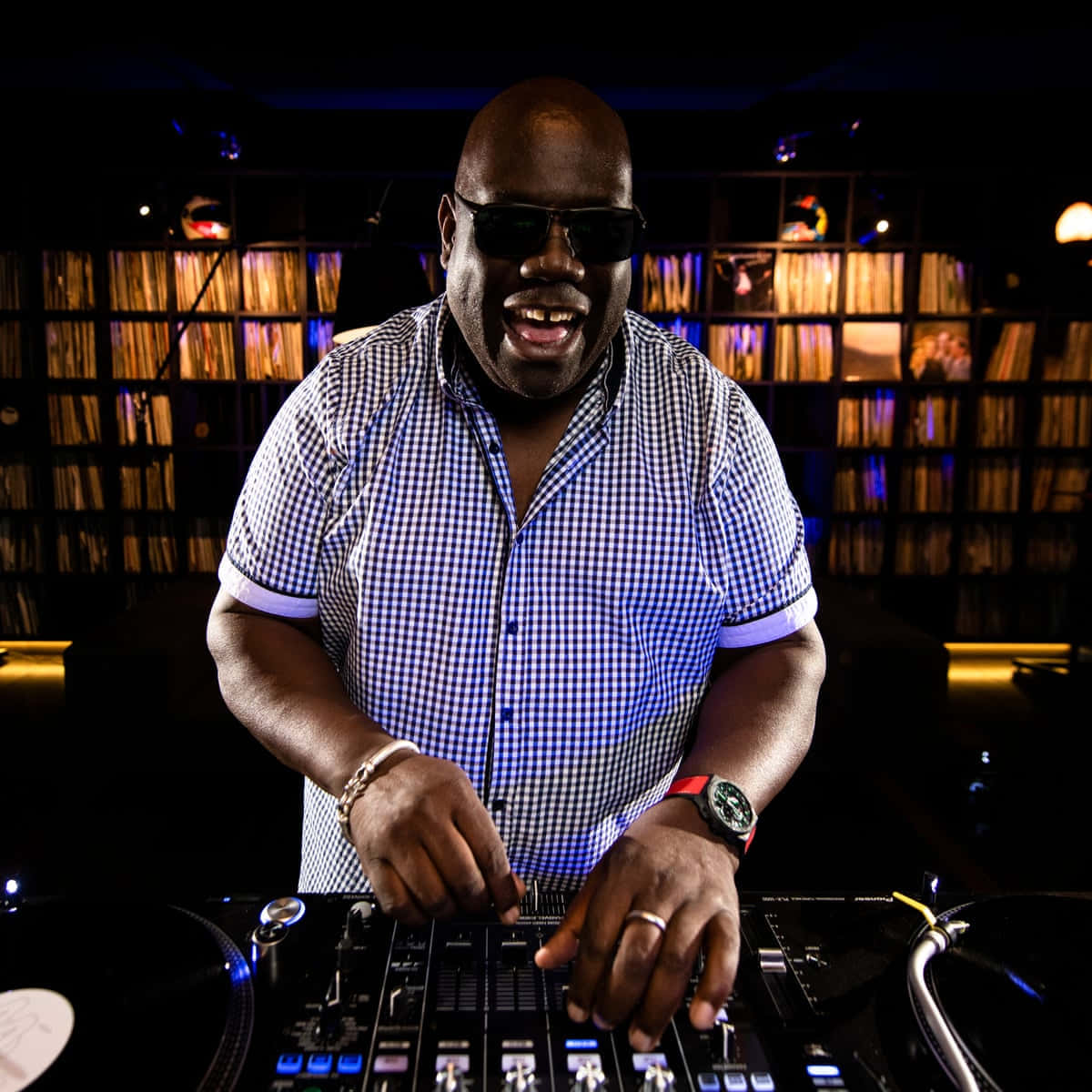 DJ 桌布 壁紙 — 一位面帶微笑的DJ穿著格仔襯衫，在昏暗的俱樂部燈光下，於帶有發光控制器的唱盤上混音。