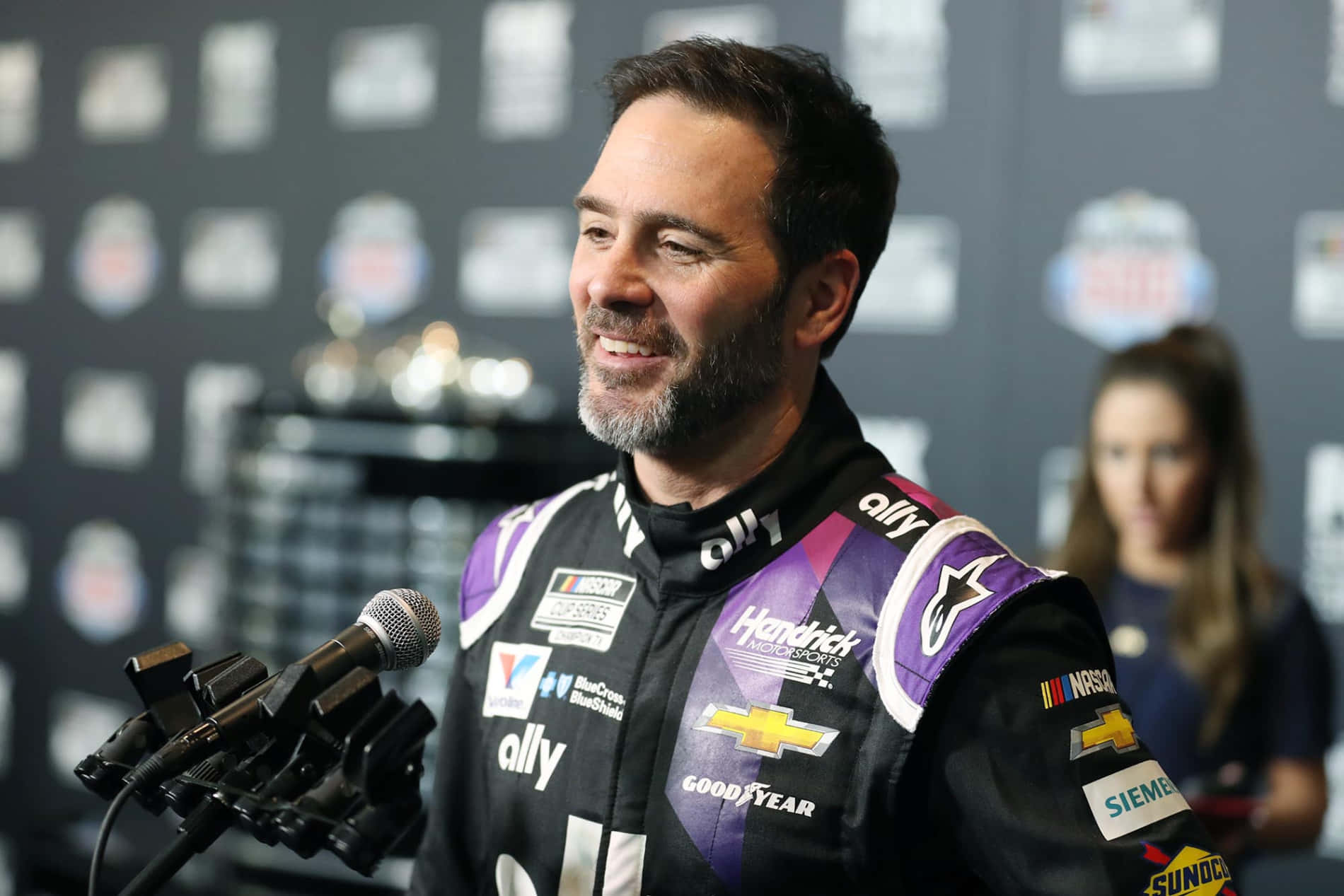 Jimmie Johnson 桌布 壁紙 — 一位面帶微笑的NASCAR車手穿著紫色和黑色的賽車服，在記者會上對著面前的麥克風發言。