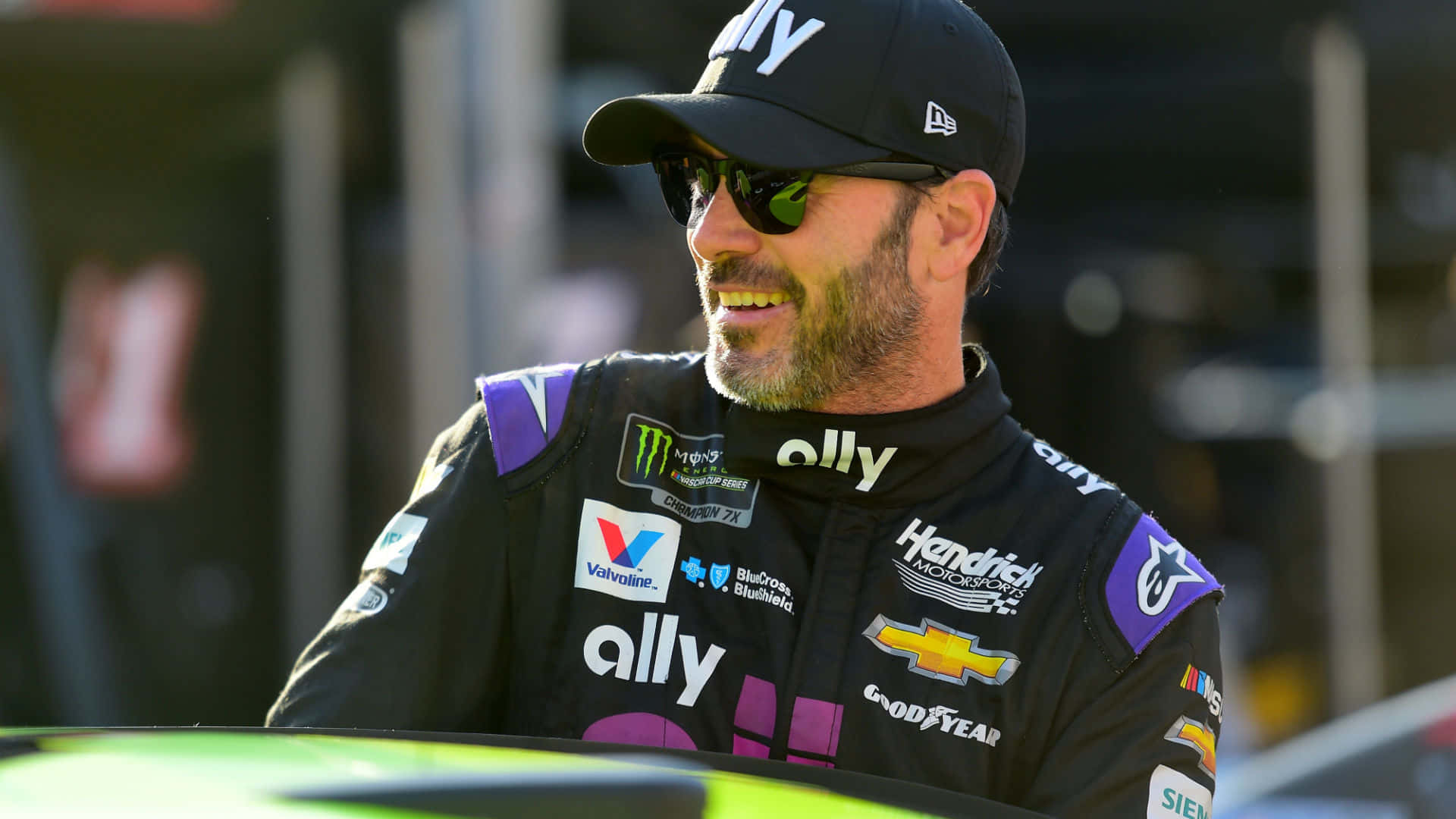 Jimmie Johnson 桌布 壁紙 — 一位面帶微笑的NASCAR車手身穿帶有贊助商標誌的黑色賽車服，靠在他的綠色賽車上。