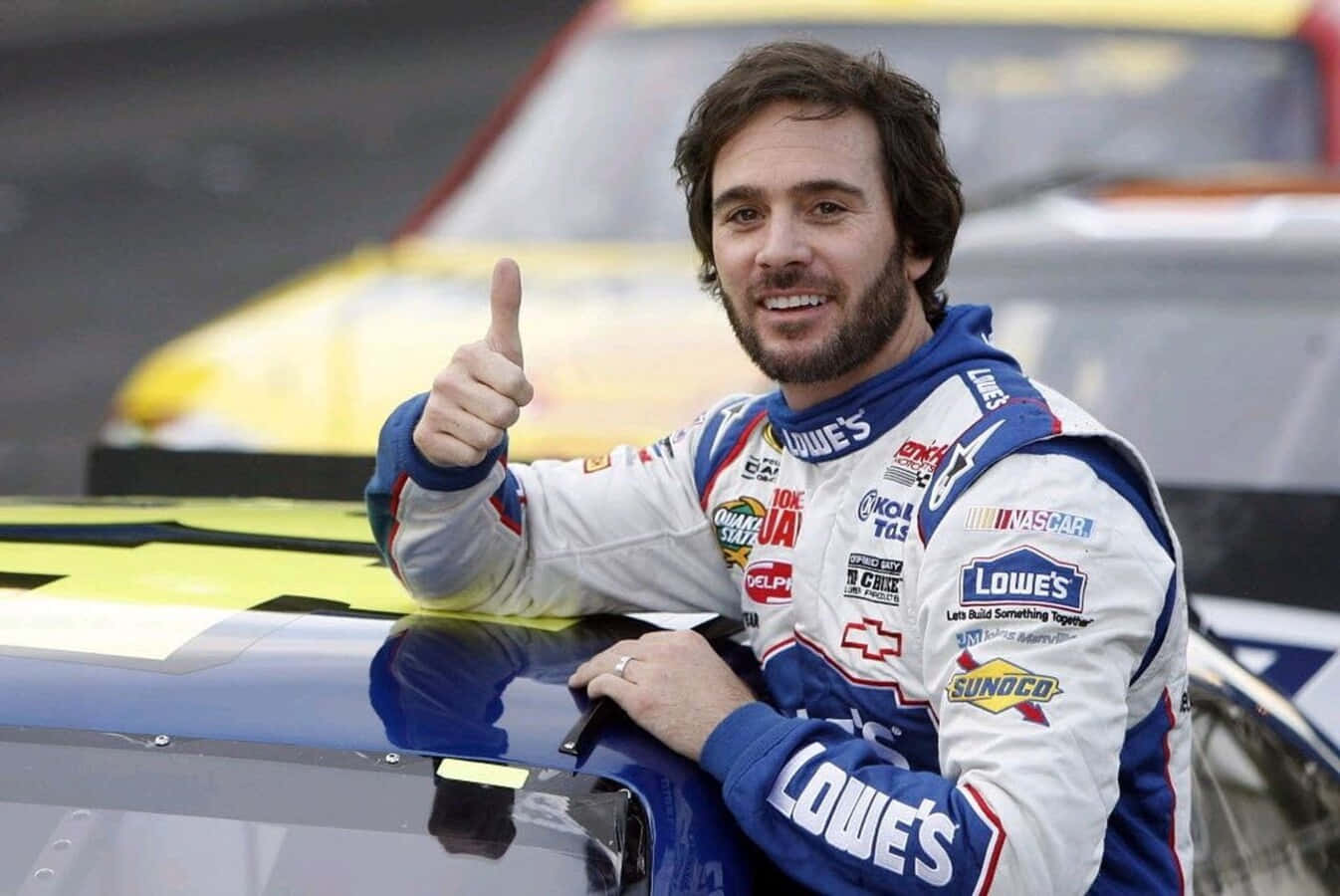 Jimmie Johnson 桌布 壁紙 — 一位面帶微笑的NASCAR車手身穿Lowe's賽車服，在他的藍色賽車旁豎起大拇指。