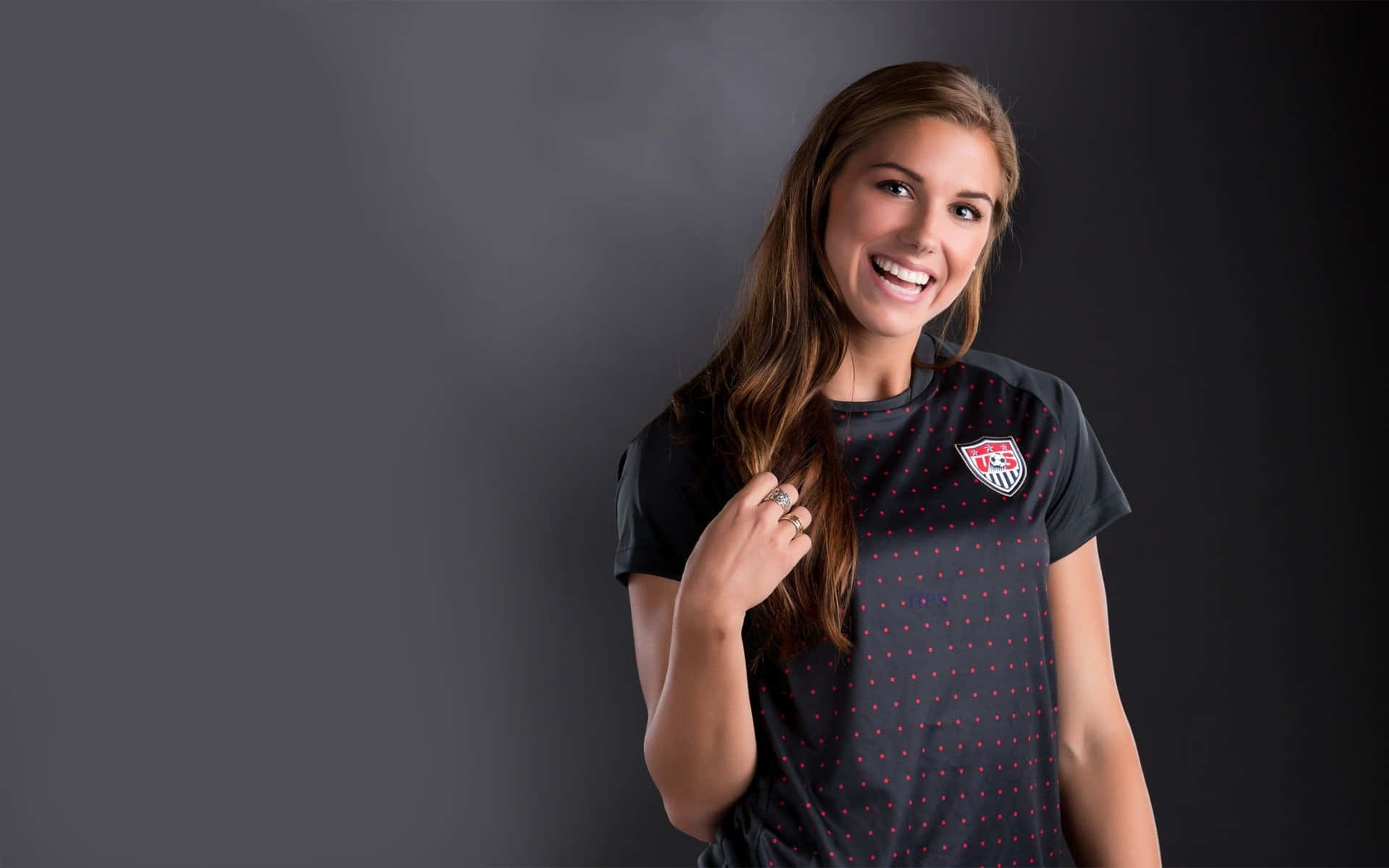 Alex Morgan 桌布 壁紙 — 一位面帶微笑的女性穿著深色圖案運動球衣，在純灰色攝影棚背景前擺姿勢。