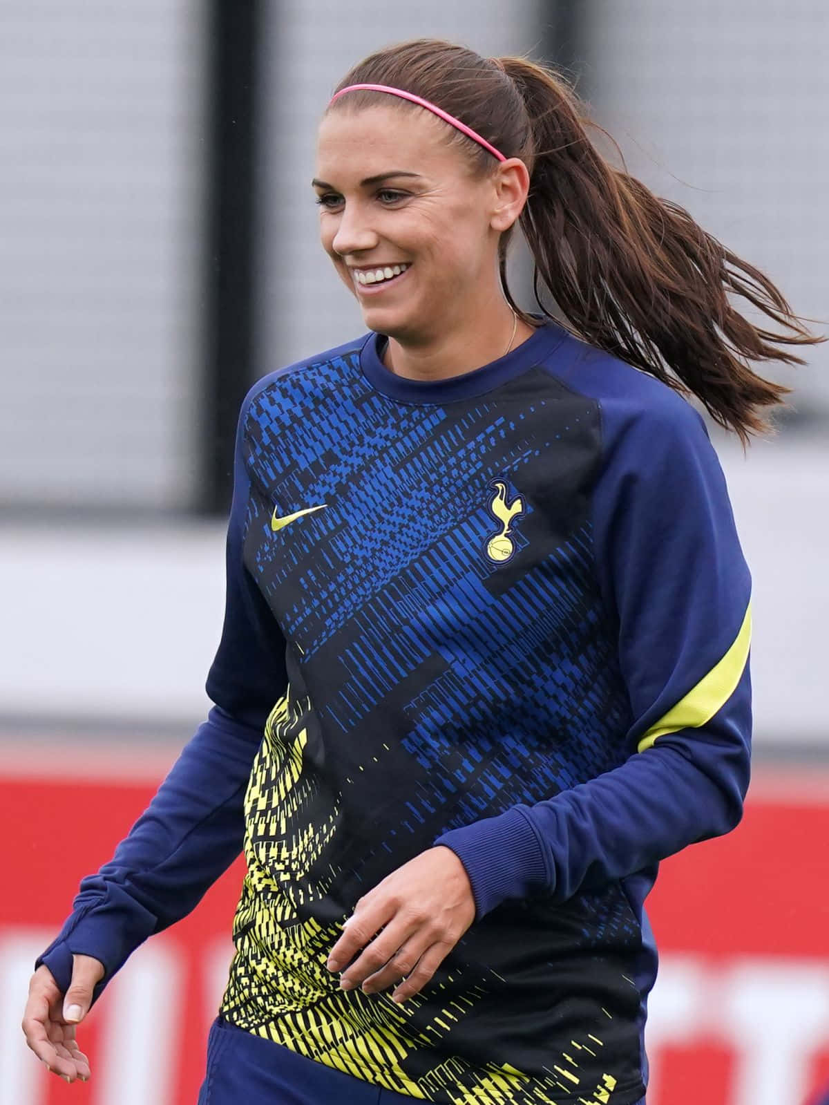 Alex Morgan 桌布 壁紙 — 一位面帶微笑的女足球員身穿藍黃色的熱刺訓練服，在風中奔跑，頭髮隨風飄揚。
