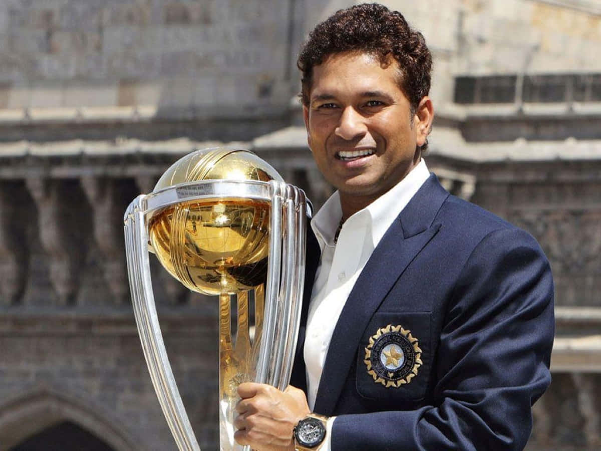 Sachin Tendulkar 桌布 壁紙 — 一位面帶微笑的板球運動員身穿海軍藍西裝外套，在歷史悠久的石砌建築背景下，手持閃閃發光的金色板球獎盃。