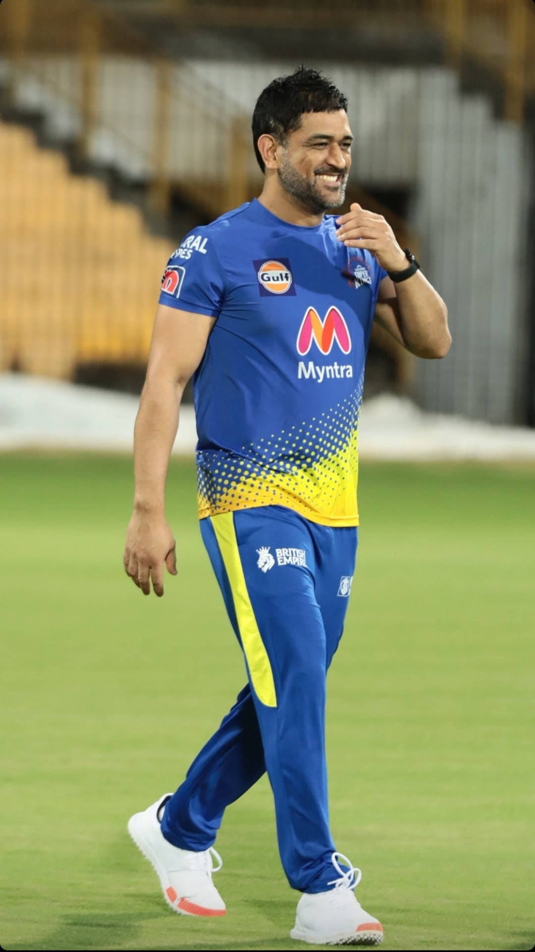 Ms Dhoni 桌布 壁紙 — 一位面帶微笑的板球運動員身穿藍黃色制服，在夜間自信地走過綠色球場。