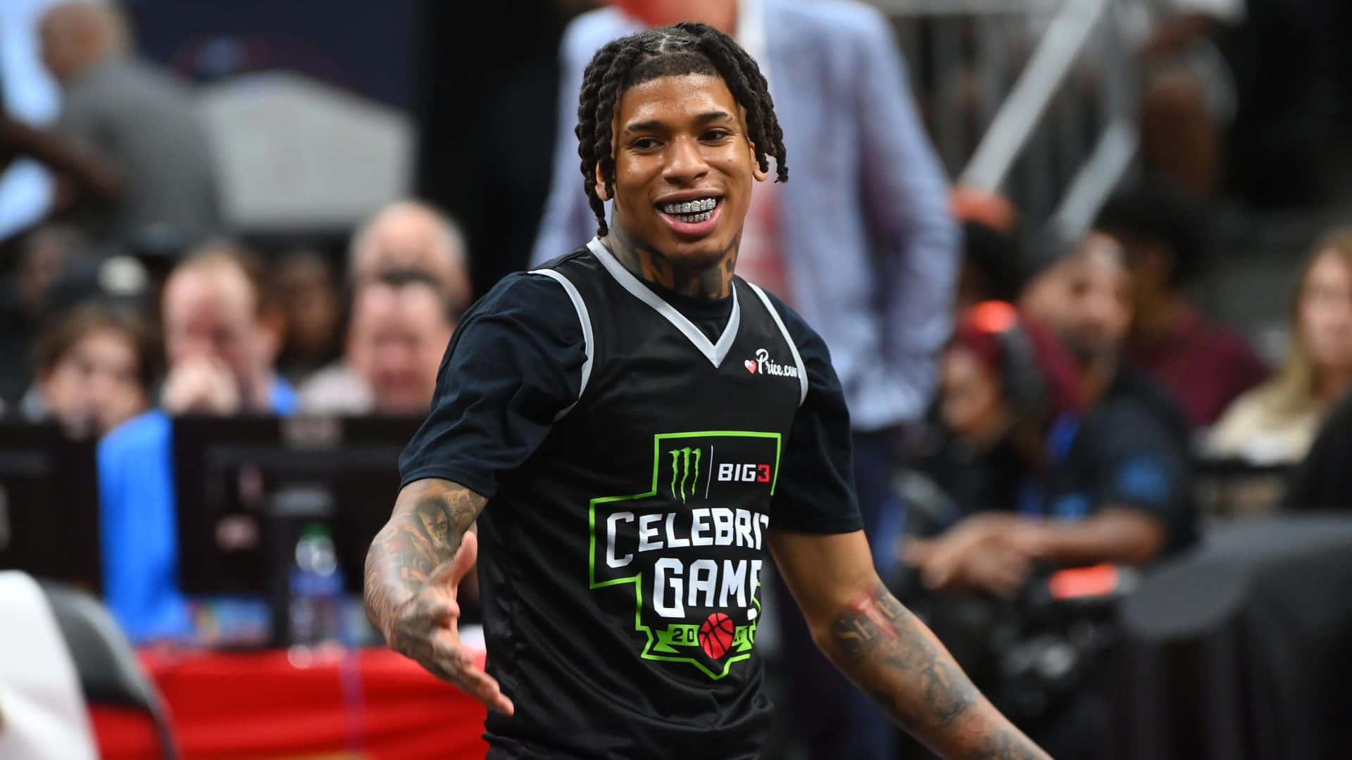 NLE Choppa 桌布 壁紙 — 一位面帶微笑的籃球運動員身穿黑色「Celebrity Game」球衣，在室內體育賽事中向人群做手勢。